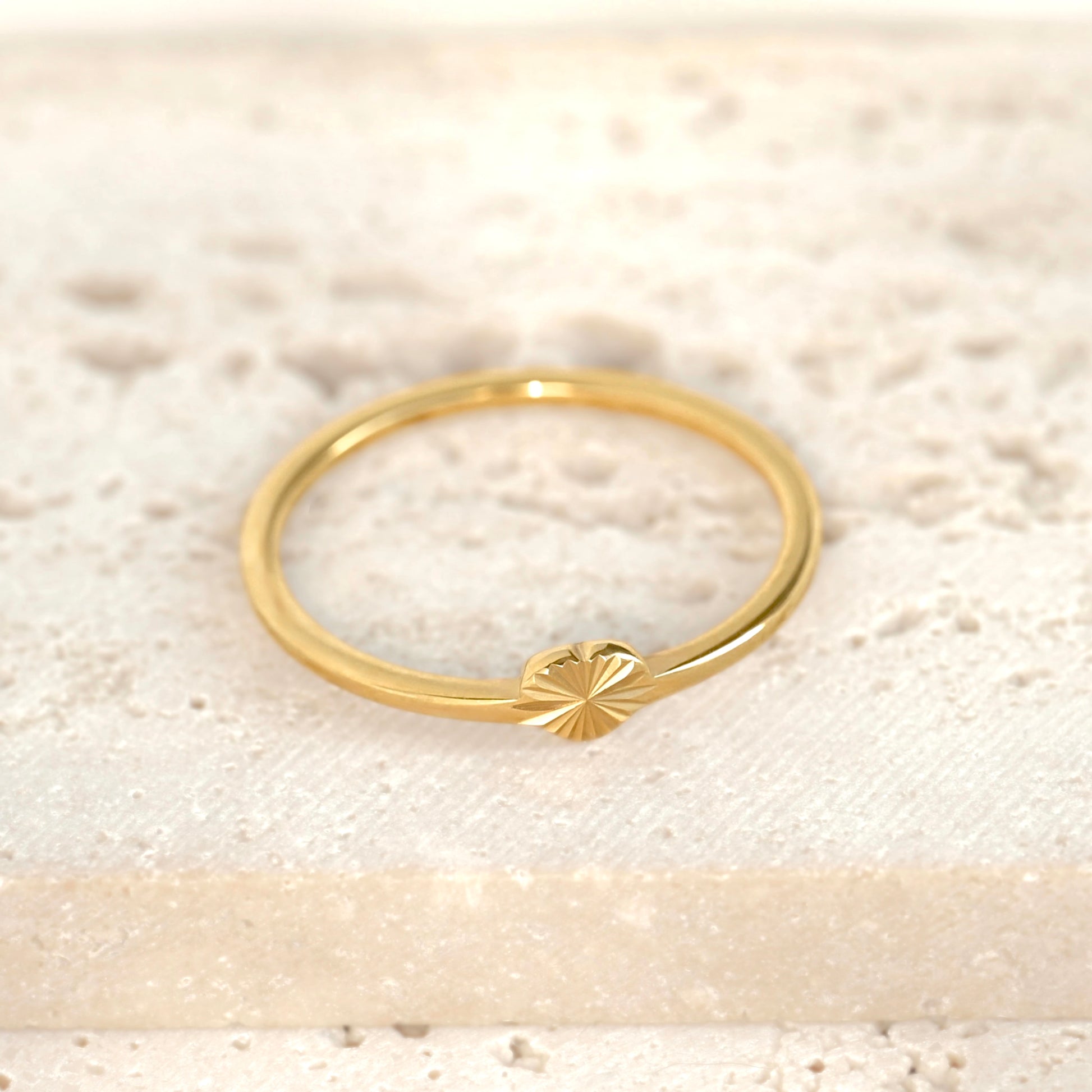 A delicate gold ring featuring a small sunburst heart detail on the front, resting on a textured, light beige stone surface.