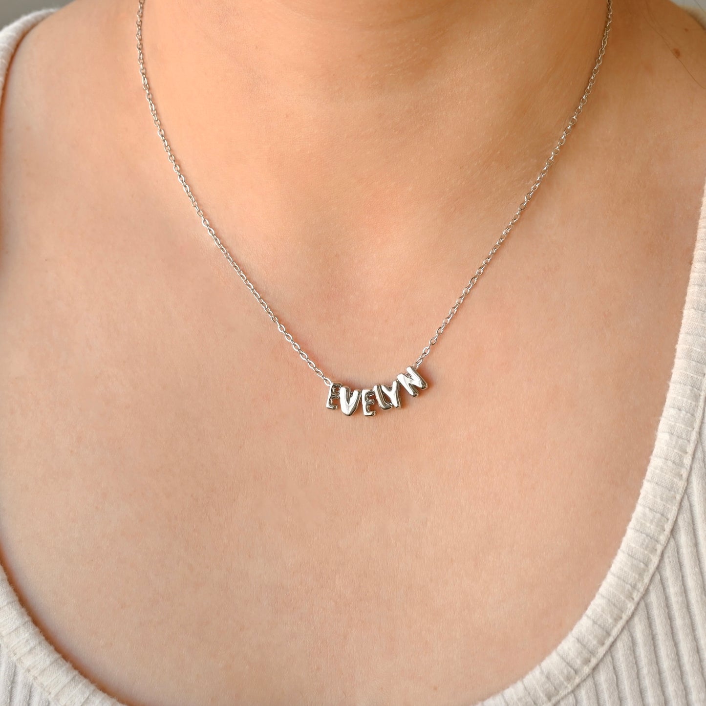 A close-up of a person wearing a delicate silver chain necklace with a personalized nameplate pendant spelling 'EvELYN' made with individual bubble initial charms