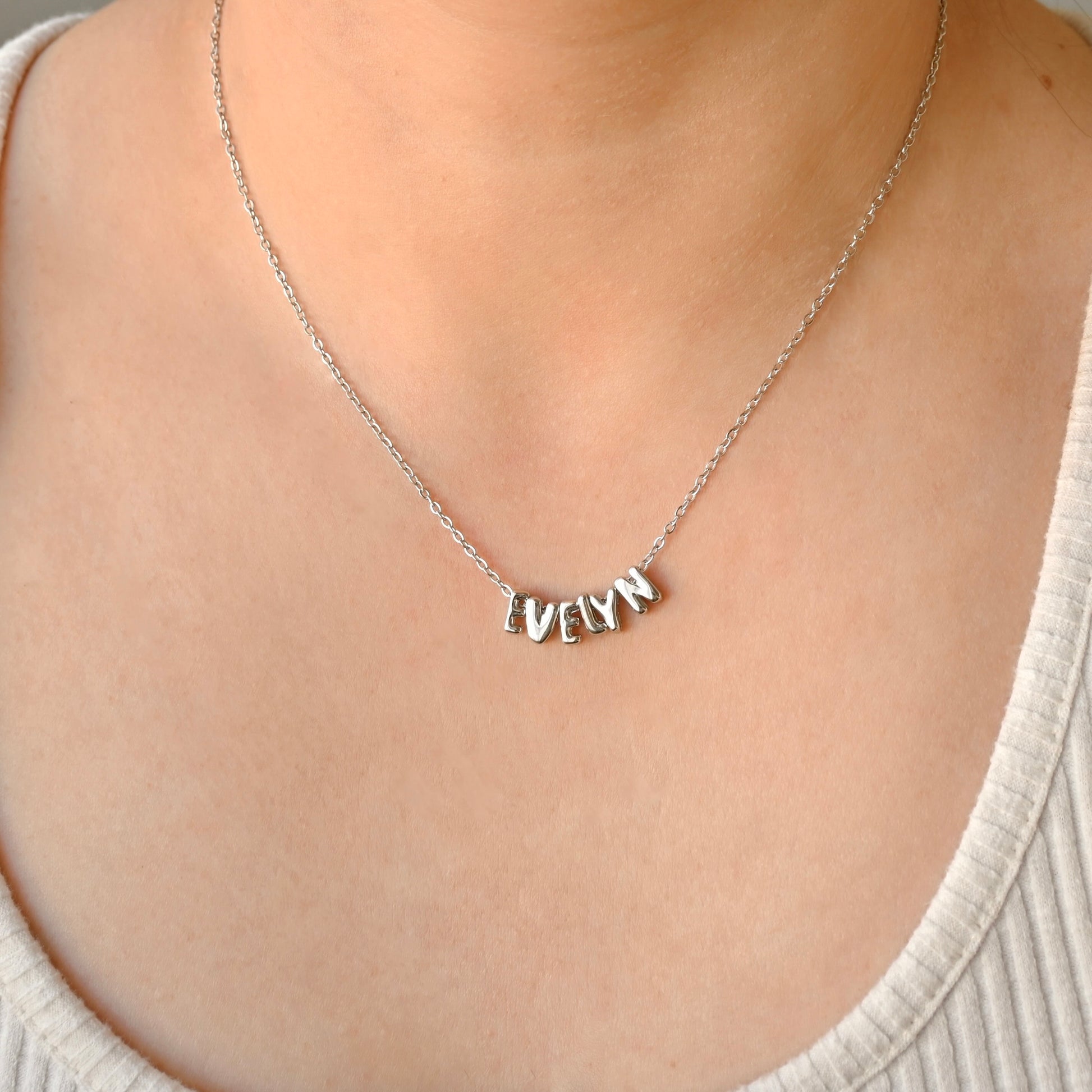 A close-up of a person wearing a delicate silver chain necklace with a personalized nameplate pendant spelling 'EvELYN' made with individual bubble initial charms