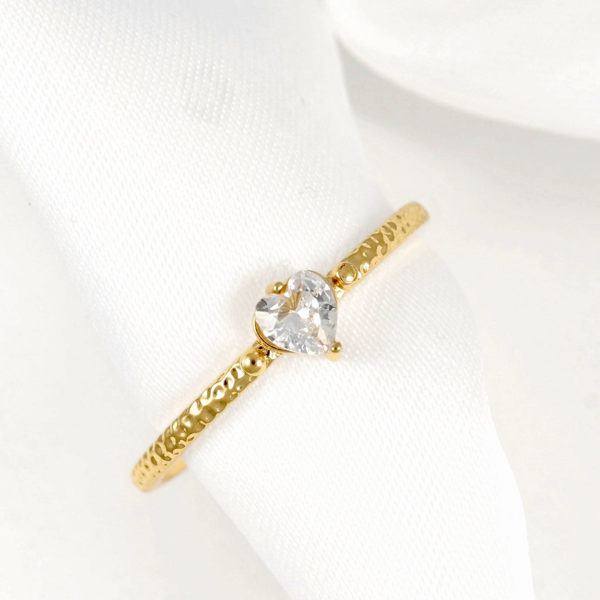 A gold ring with a textured band and a clear heart-shaped gemstone, displayed on a white fabric ring holder.