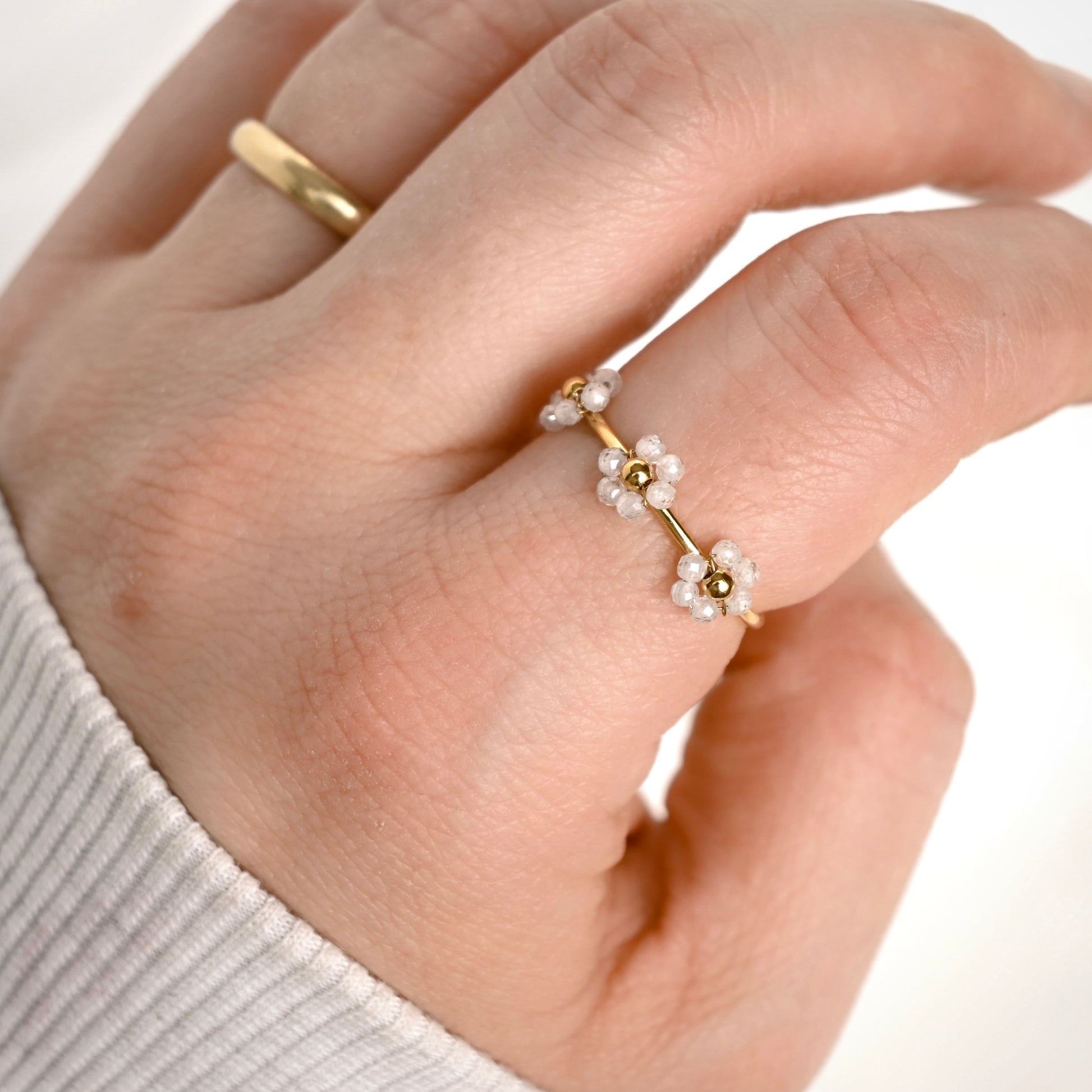 A close-up of a hand wearing a dainty gold ring adorned with small white beaded flowers, with a simple gold band on another finger, set against a clean white background.