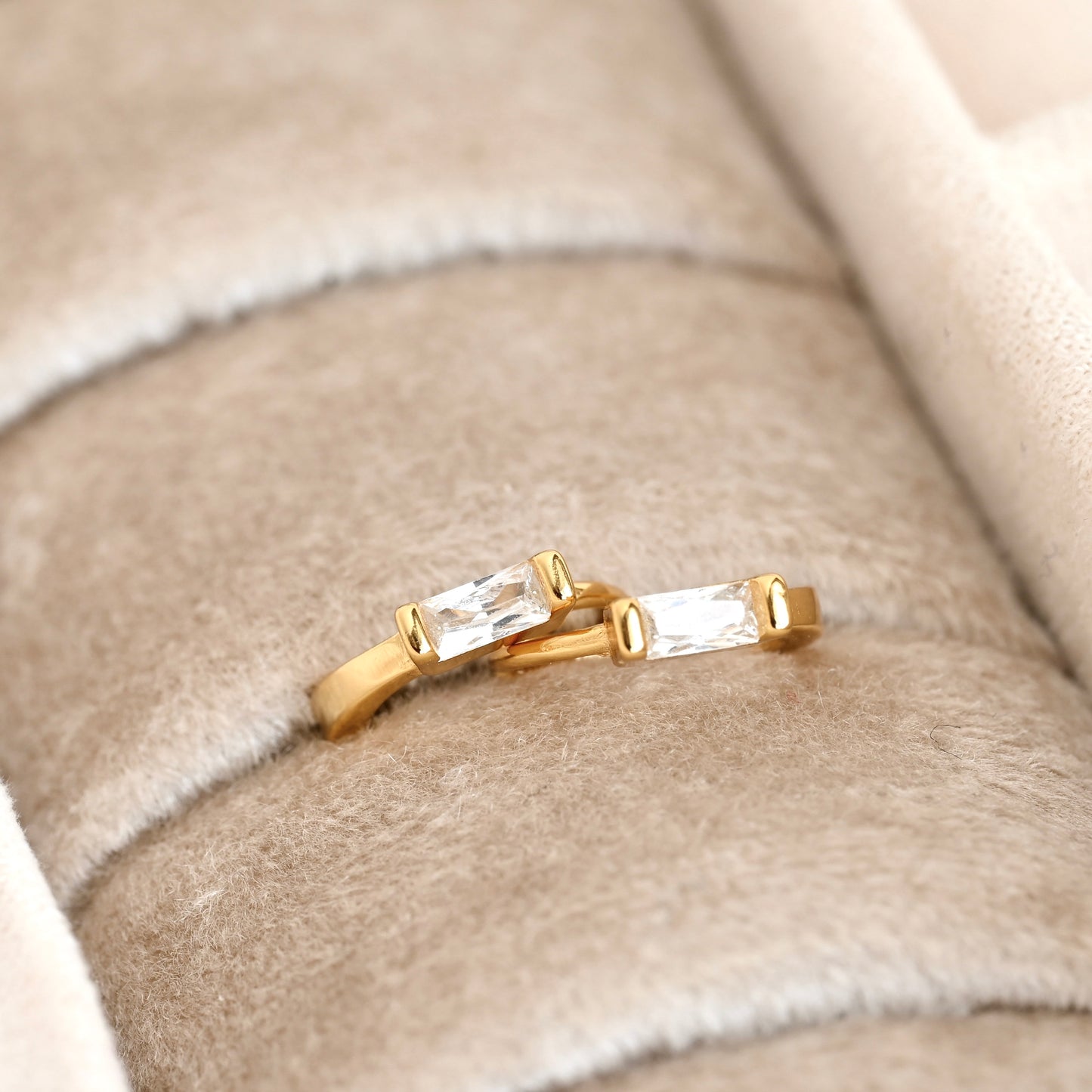 Two gold rings with rectangular clear gemstones, displayed on a beige velvet ring box.