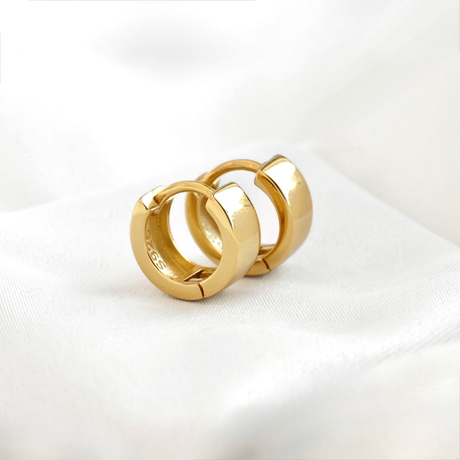 A pair of polished small gold huggie hoop earrings resting on a soft, white textured fabric background.