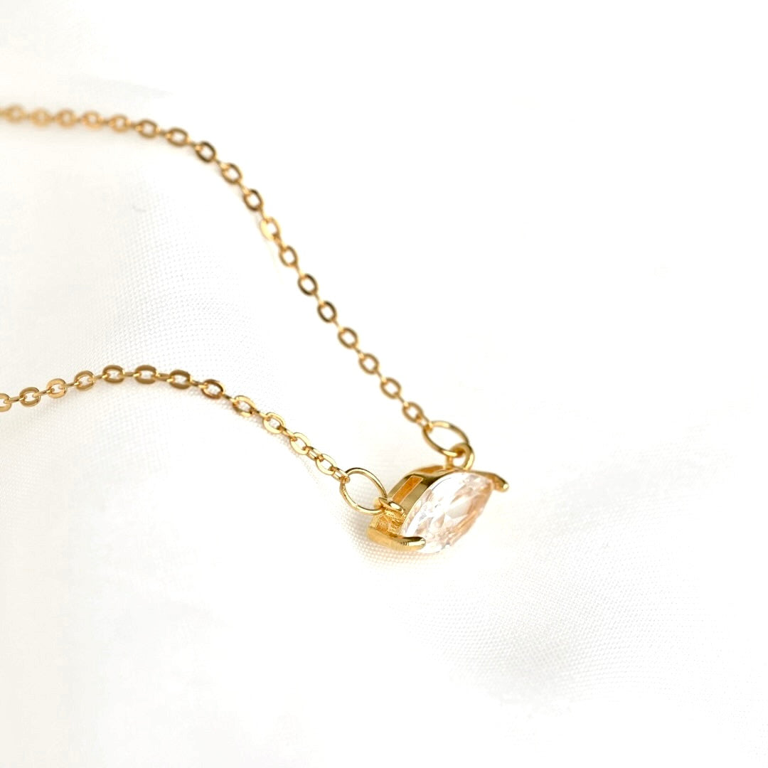 A delicate gold chain necklace with a small gold pendant holding a marquise-cut clear gemstone, displayed on a soft white fabric background.