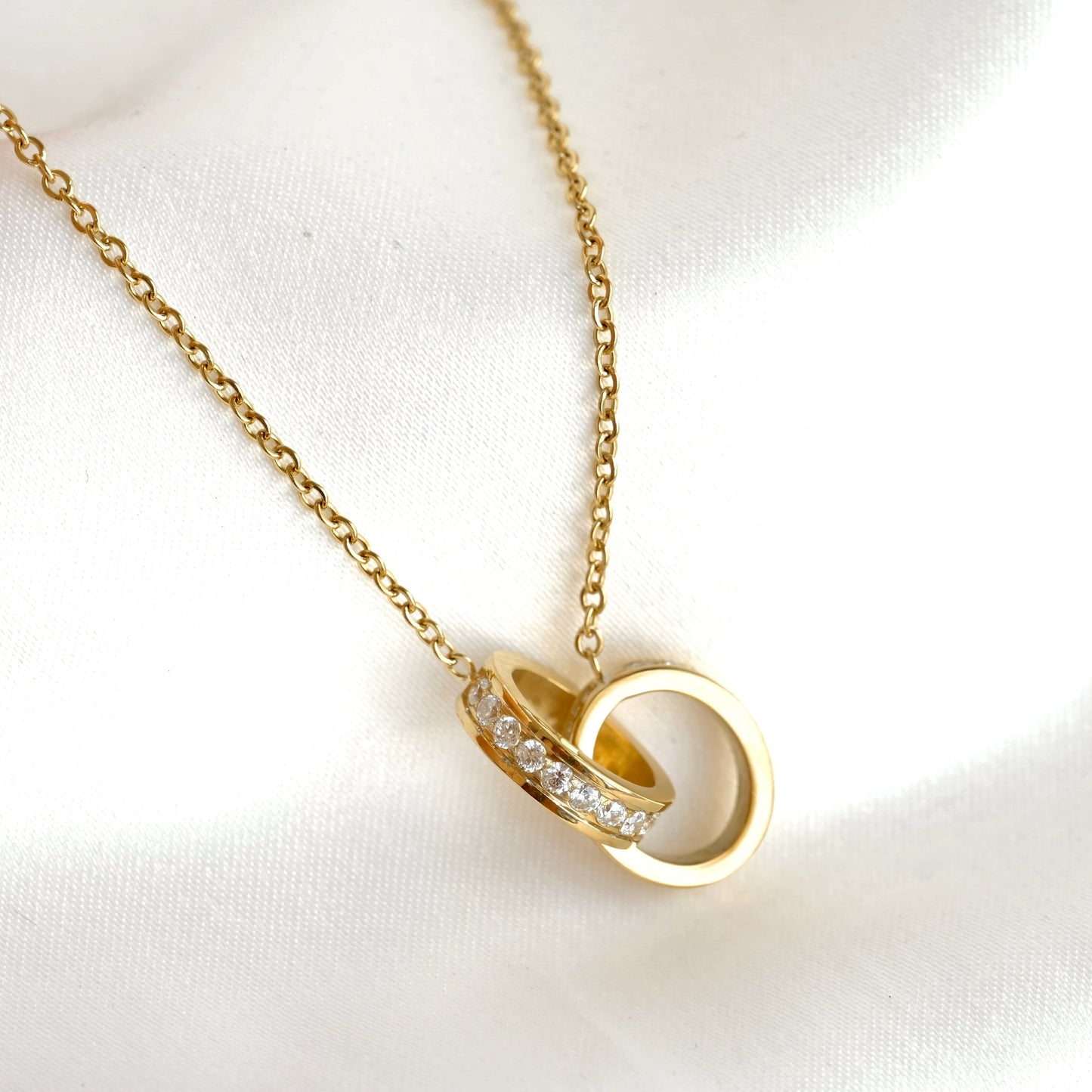 A gold chain necklace with a pendant featuring two interlocking rings with small clear CZ stones, displayed on a white satin fabric background.