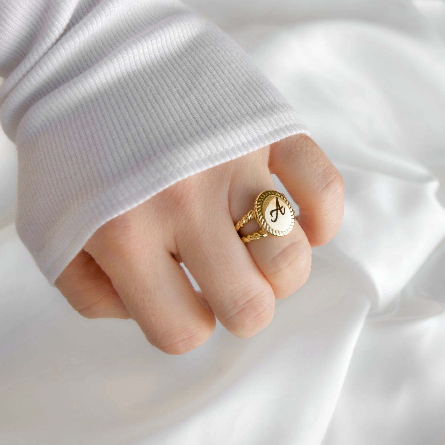 A close-up of a hand wearing a gold signet ring with a white oval face and black monogram letter 'A', styled with a white ribbed long-sleeve top against a draped white fabric background.