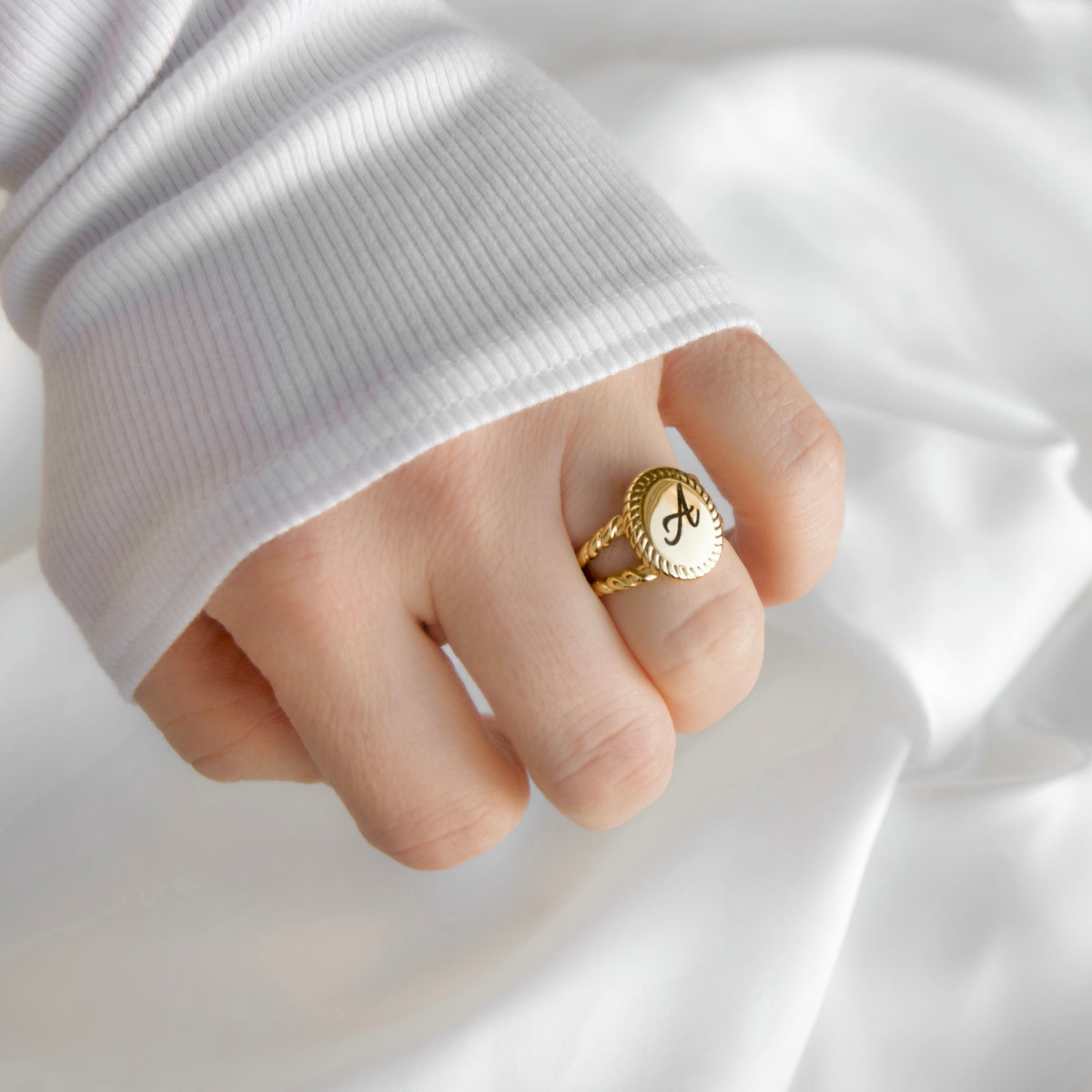 A close-up of a hand wearing a gold signet ring with a white oval face and black monogram letter 'A', styled with a white ribbed long-sleeve top against a draped white fabric background.