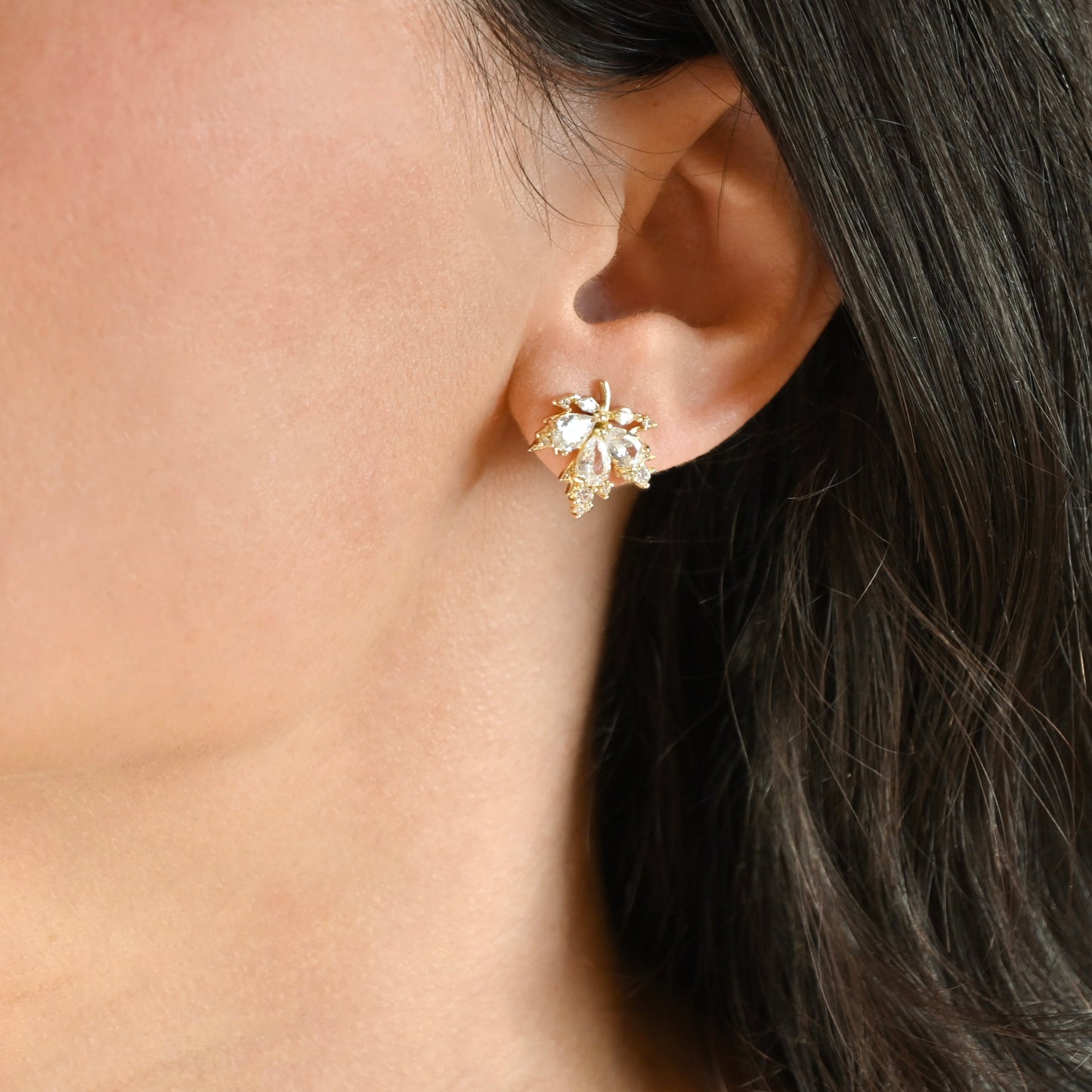 A close-up of a model's ear wearing a gold-toned leaf-shaped stud earring adorned with clear crystals.