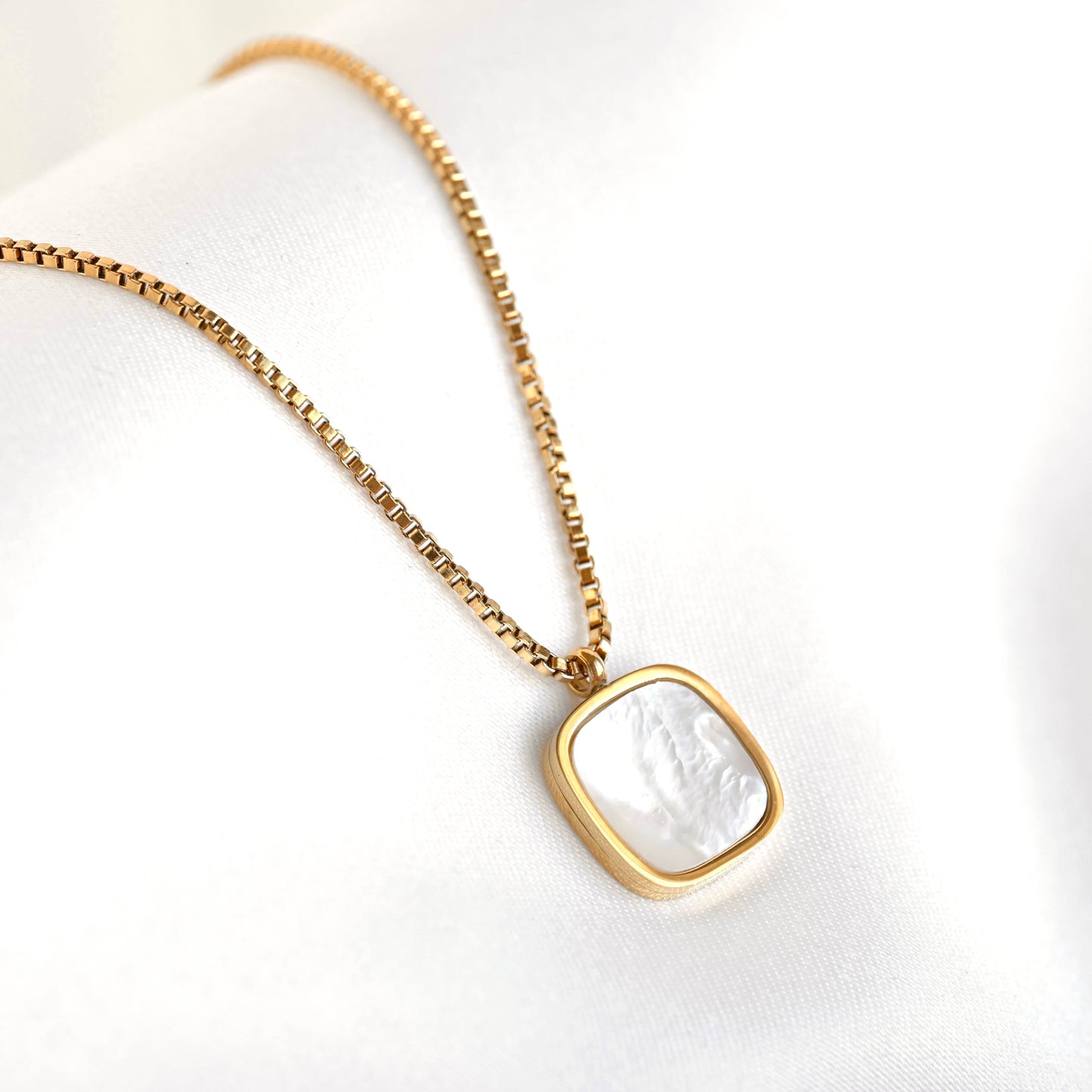 A gold chain necklace with a square bezel pendant featuring a white mother-of-pearl inlay, displayed on a soft white fabric background.