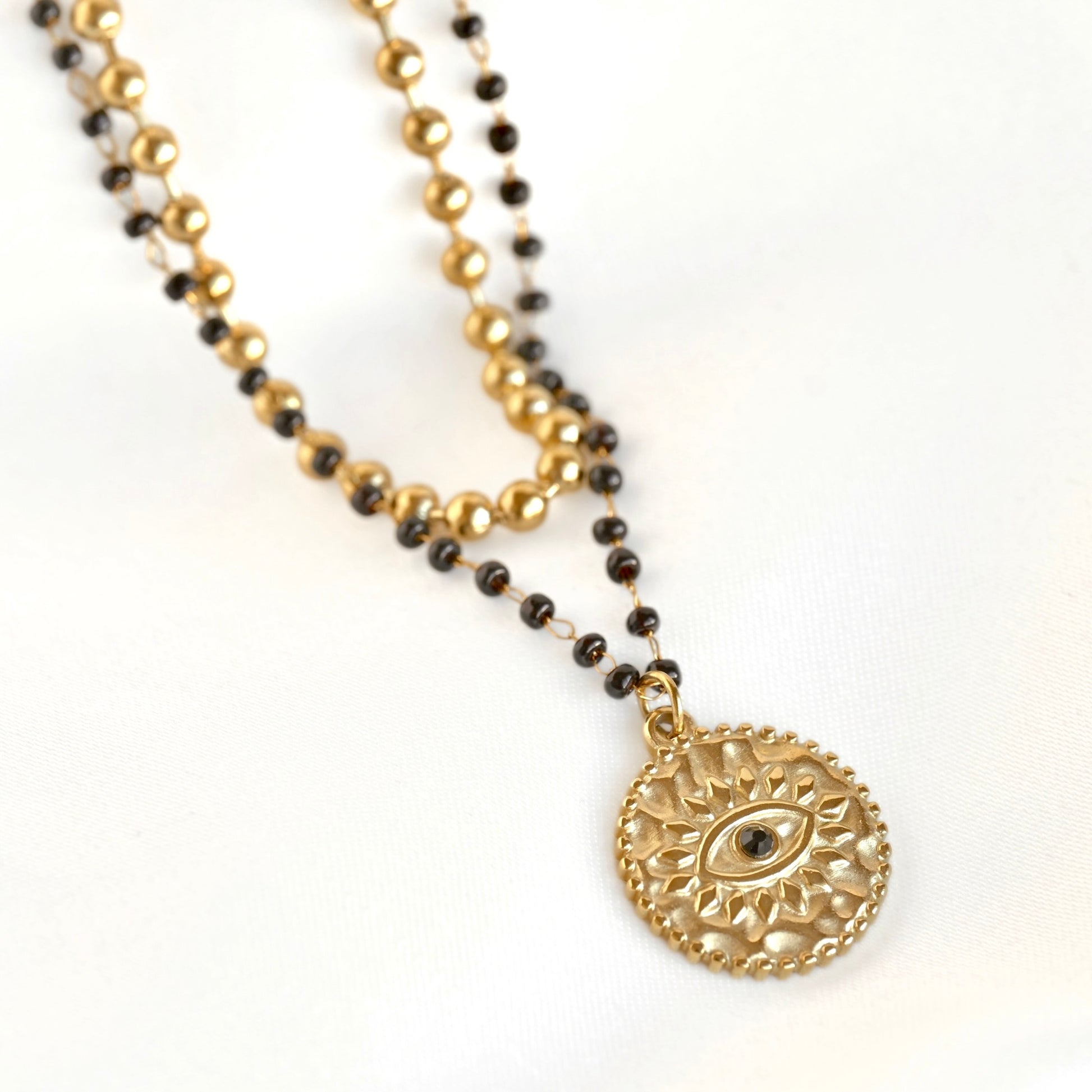 A double-strand beaded necklace featuring a gold-tone circular pendant with an embossed sun-and-eye design, paired with a secondary strand of small black and gold beads, shown on a white background.
