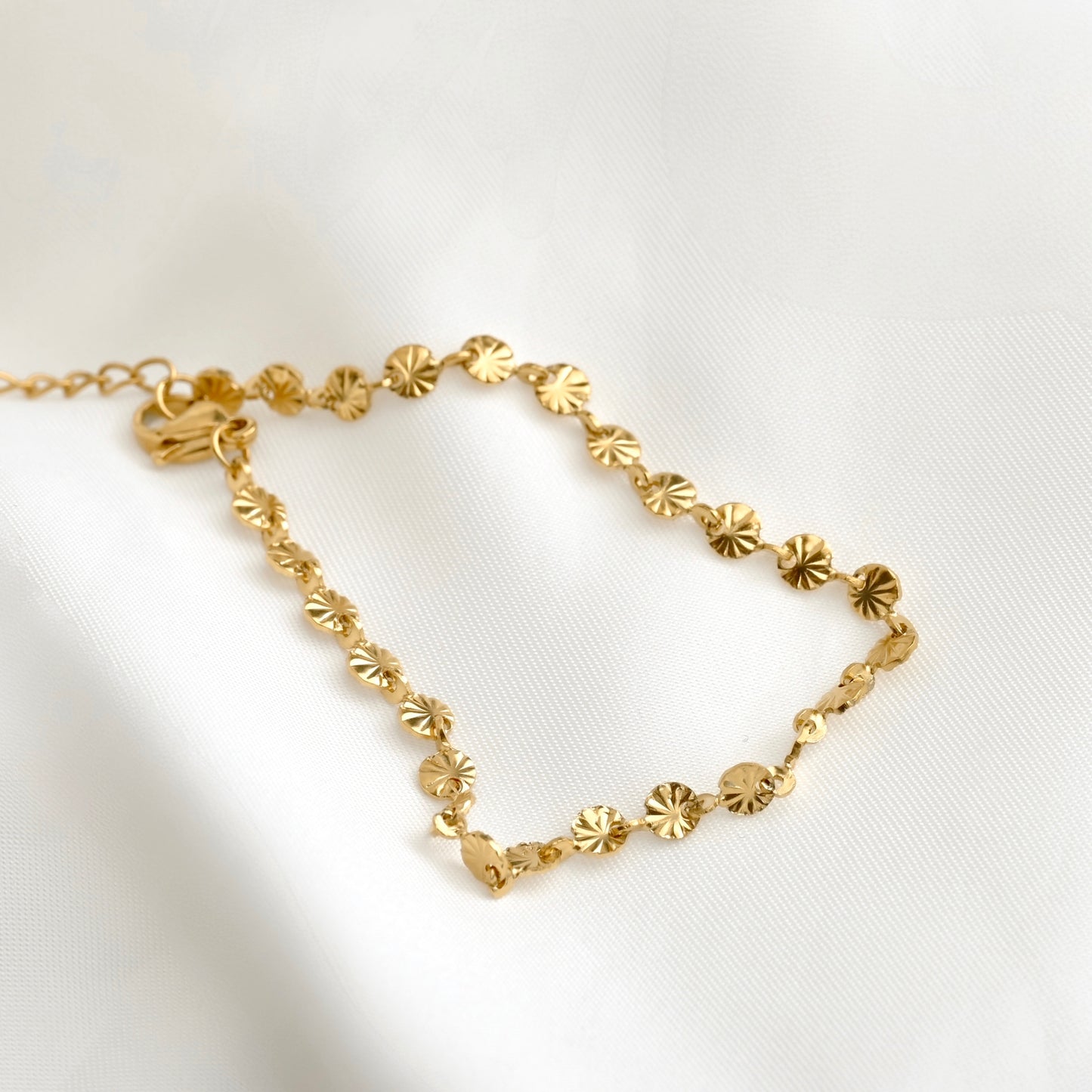 A delicate gold chain bracelet featuring small, starburst disc charms, displayed on a soft white fabric background.