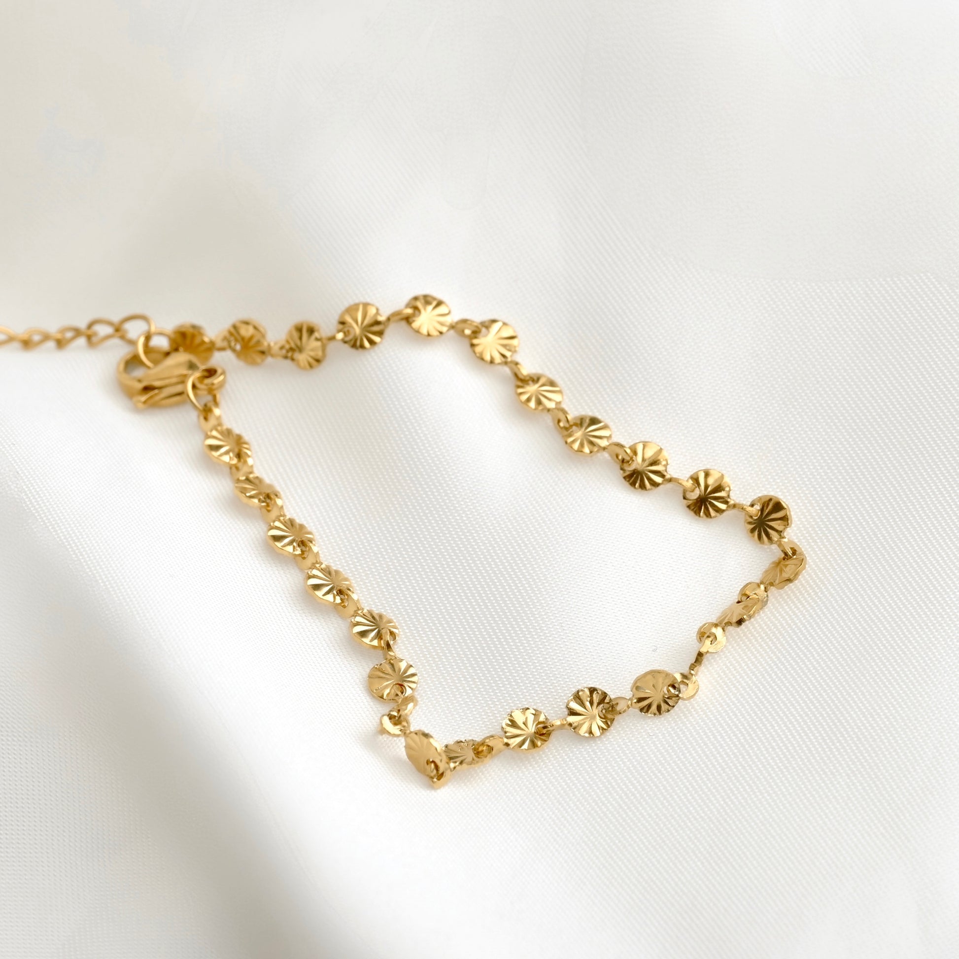 A delicate gold chain bracelet featuring small, starburst disc charms, displayed on a soft white fabric background.