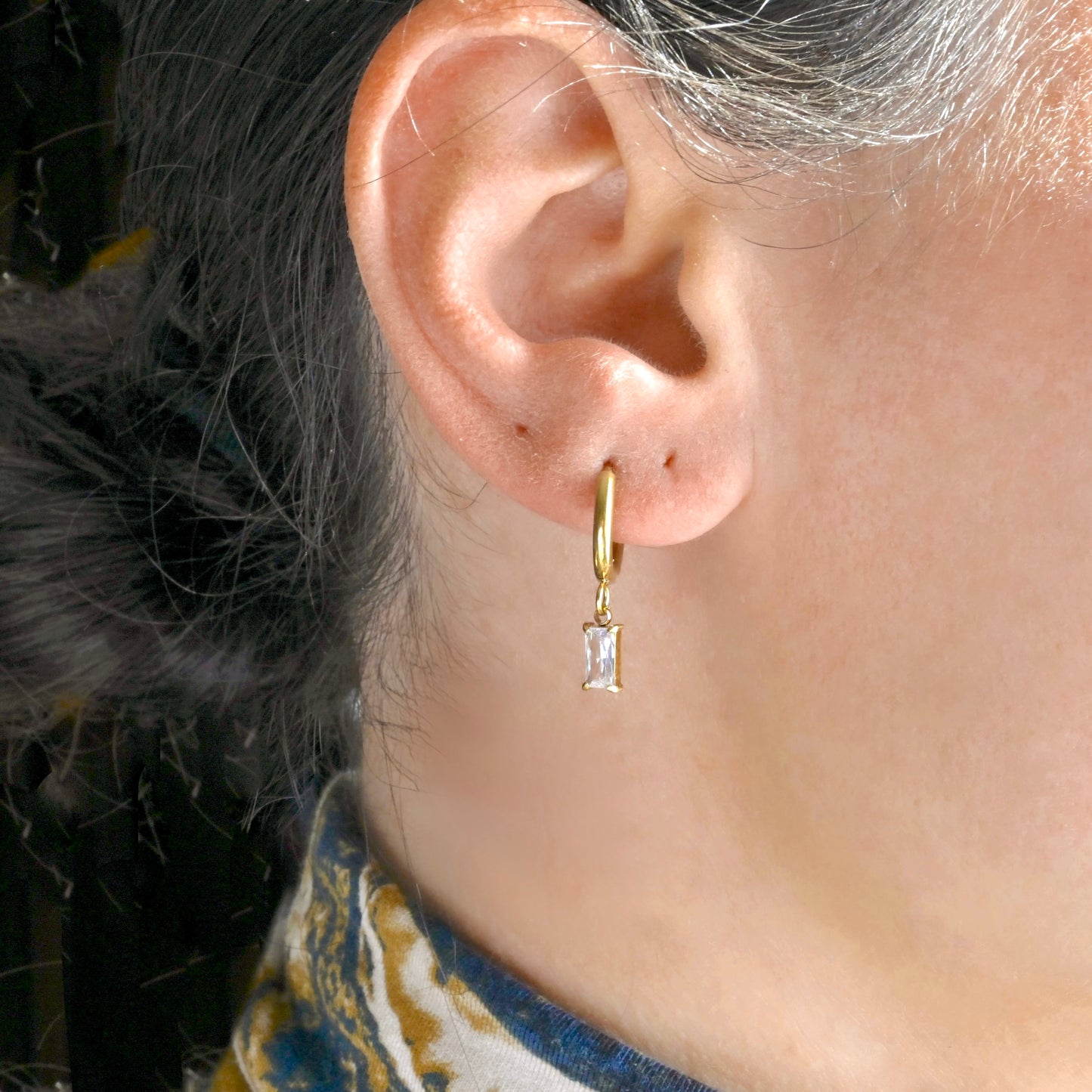 Close-up of an ear wearing a baguette charm gold hoop earring with a clear CZ.