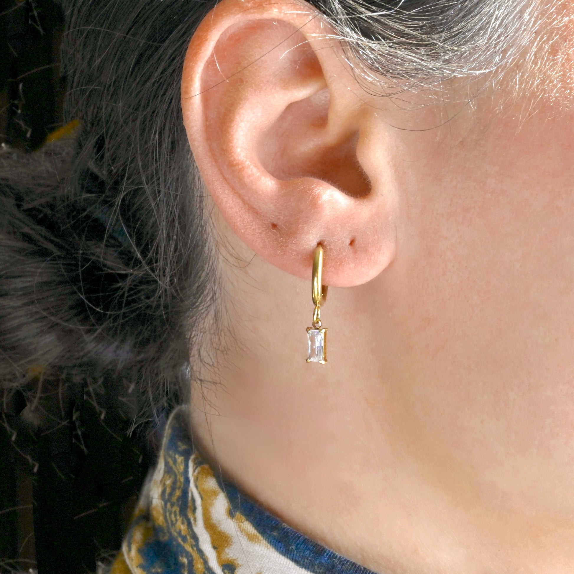 Close-up of an ear wearing a baguette charm gold hoop earring with a clear CZ.