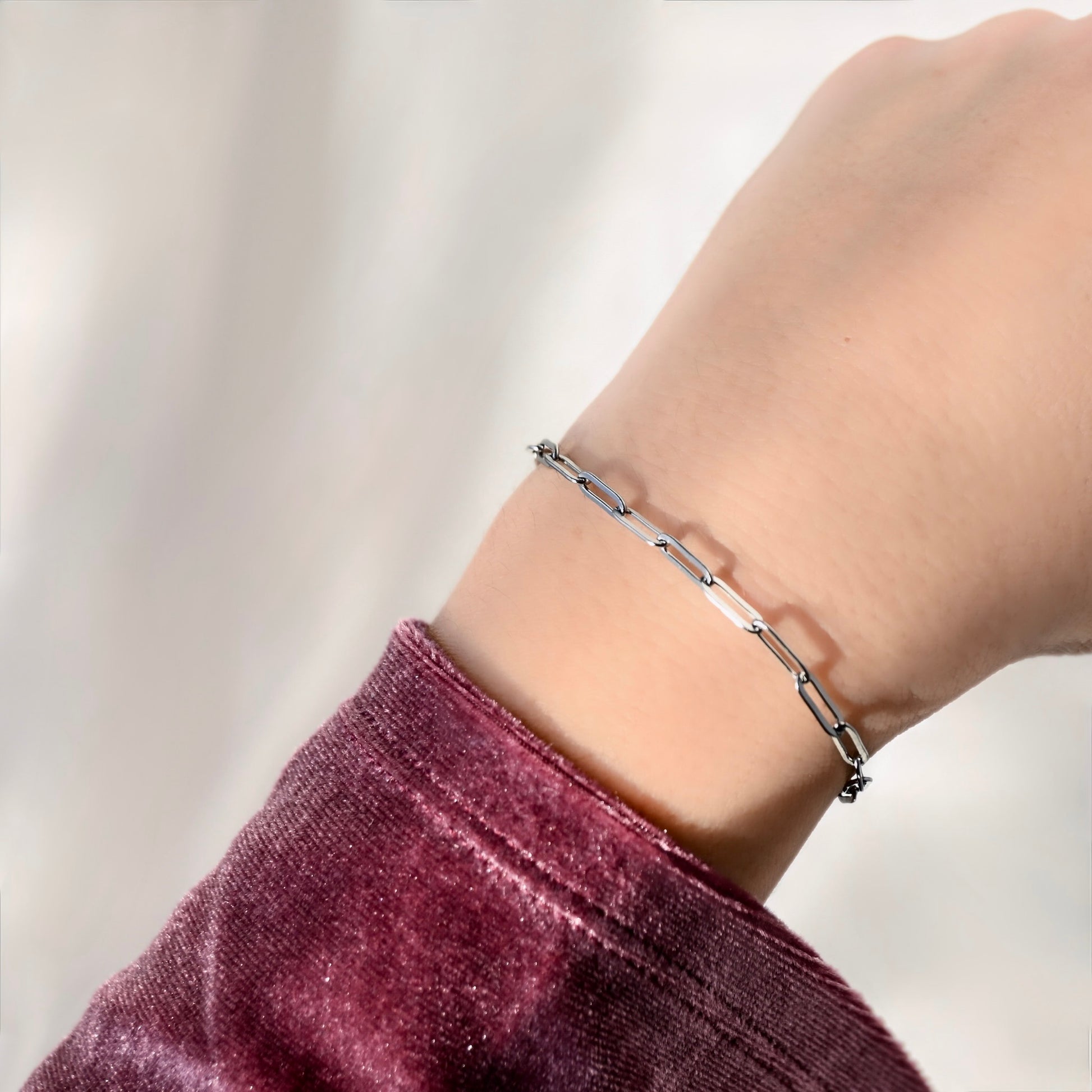 A close-up of a delicate silver chain bracelet worn on a person's wrist, paired with a deep plum velvet sleeve against a soft, light background.