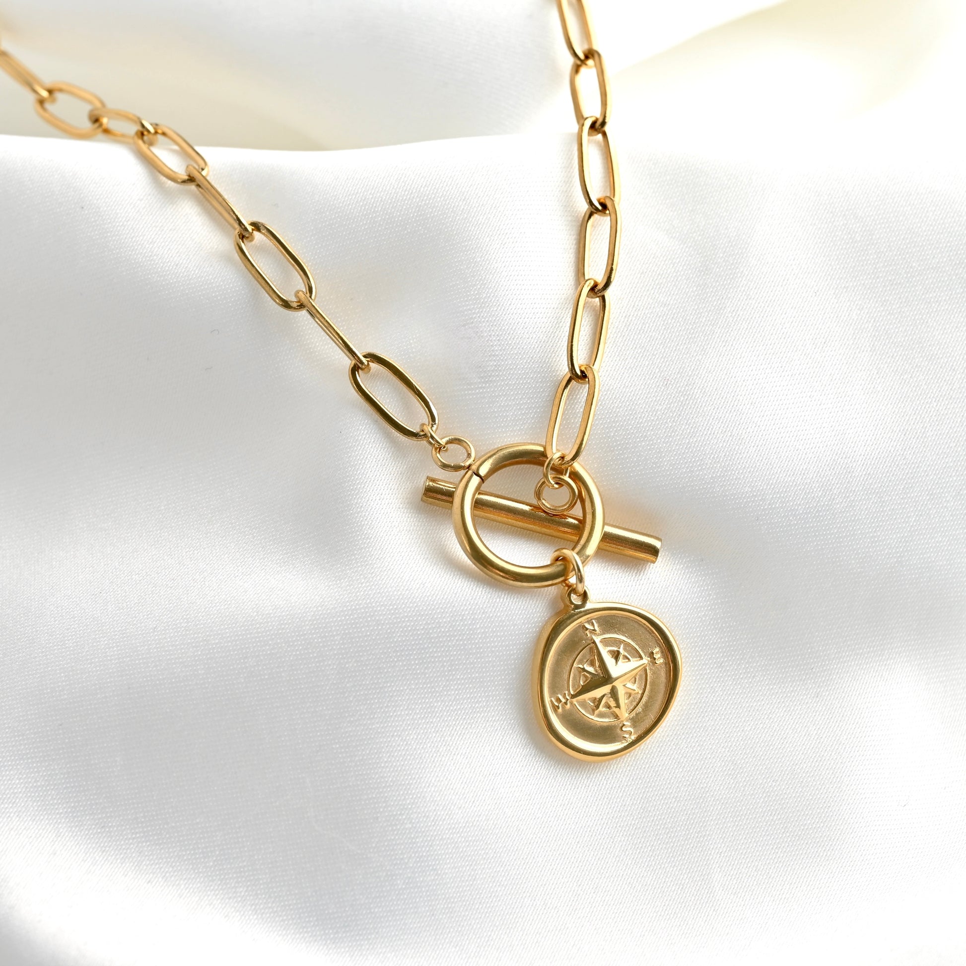 A gold-toned paperclip chain necklace with a toggle clasp and a dangling compass coin charm, displayed on a white satin fabric background.