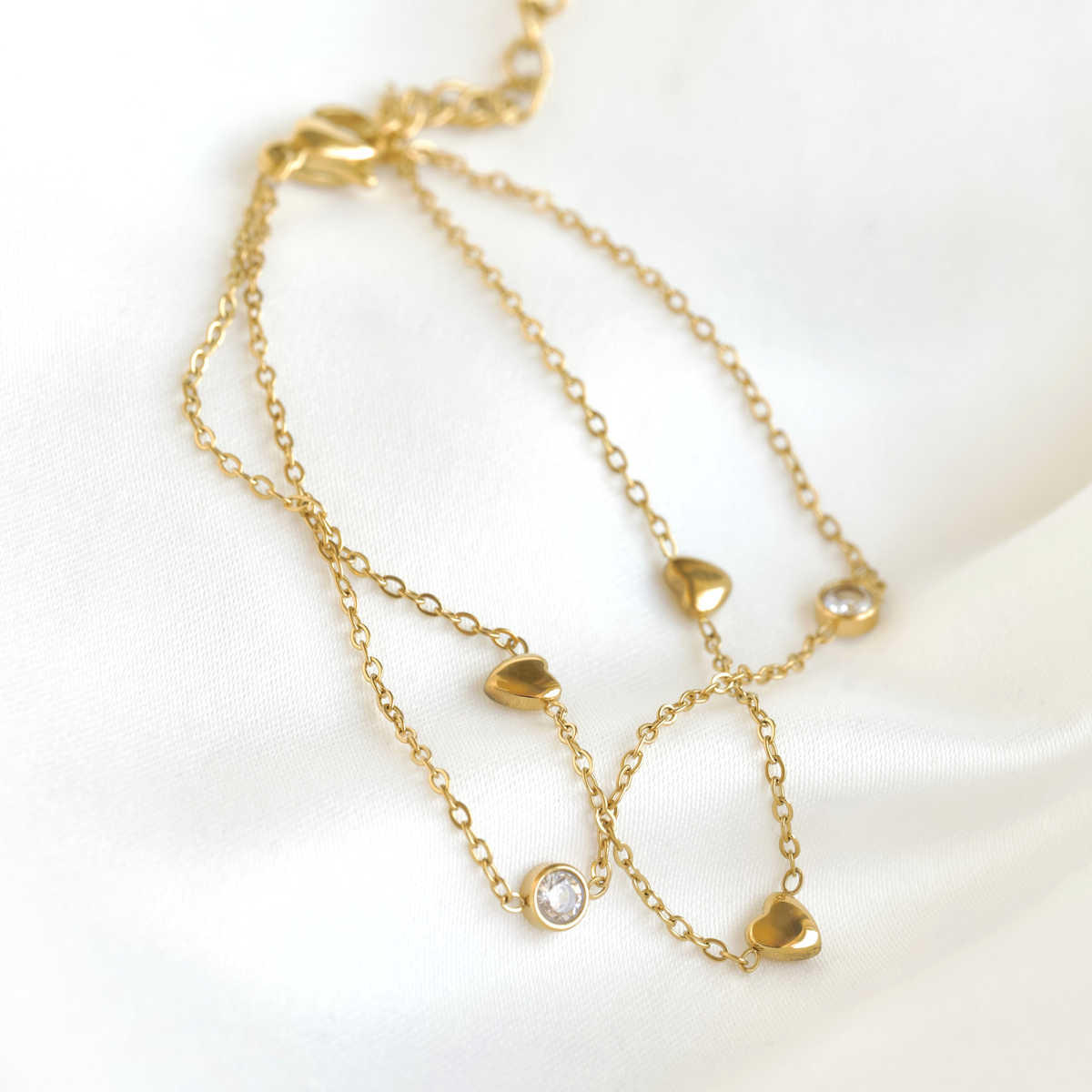 A delicate gold double-strand bracelet featuring small heart charms, one of which is adorned with a clear round gemstone, displayed on a soft white satin fabric.