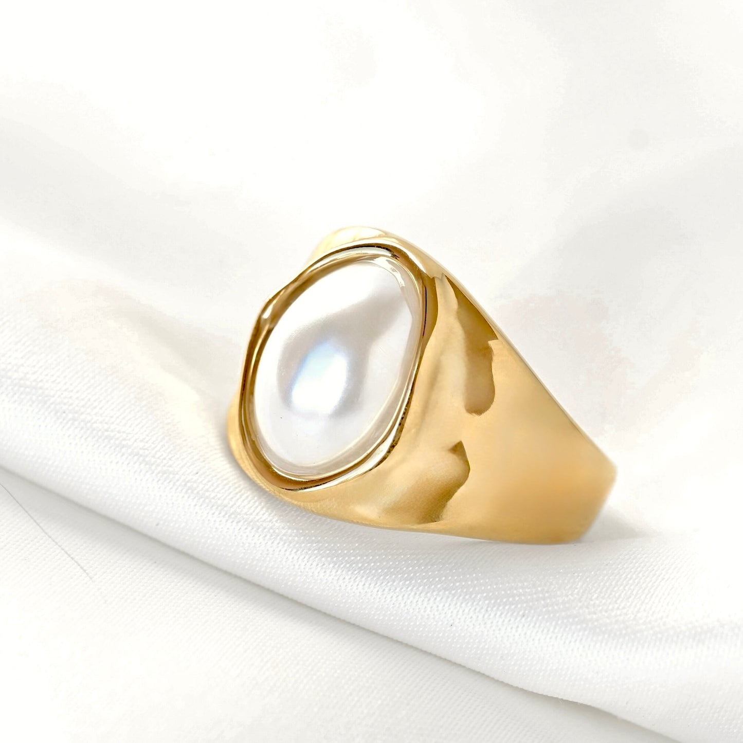 A gold signet-style ring with a large, irregular-shaped white pearl bezel-set on a soft white fabric background.