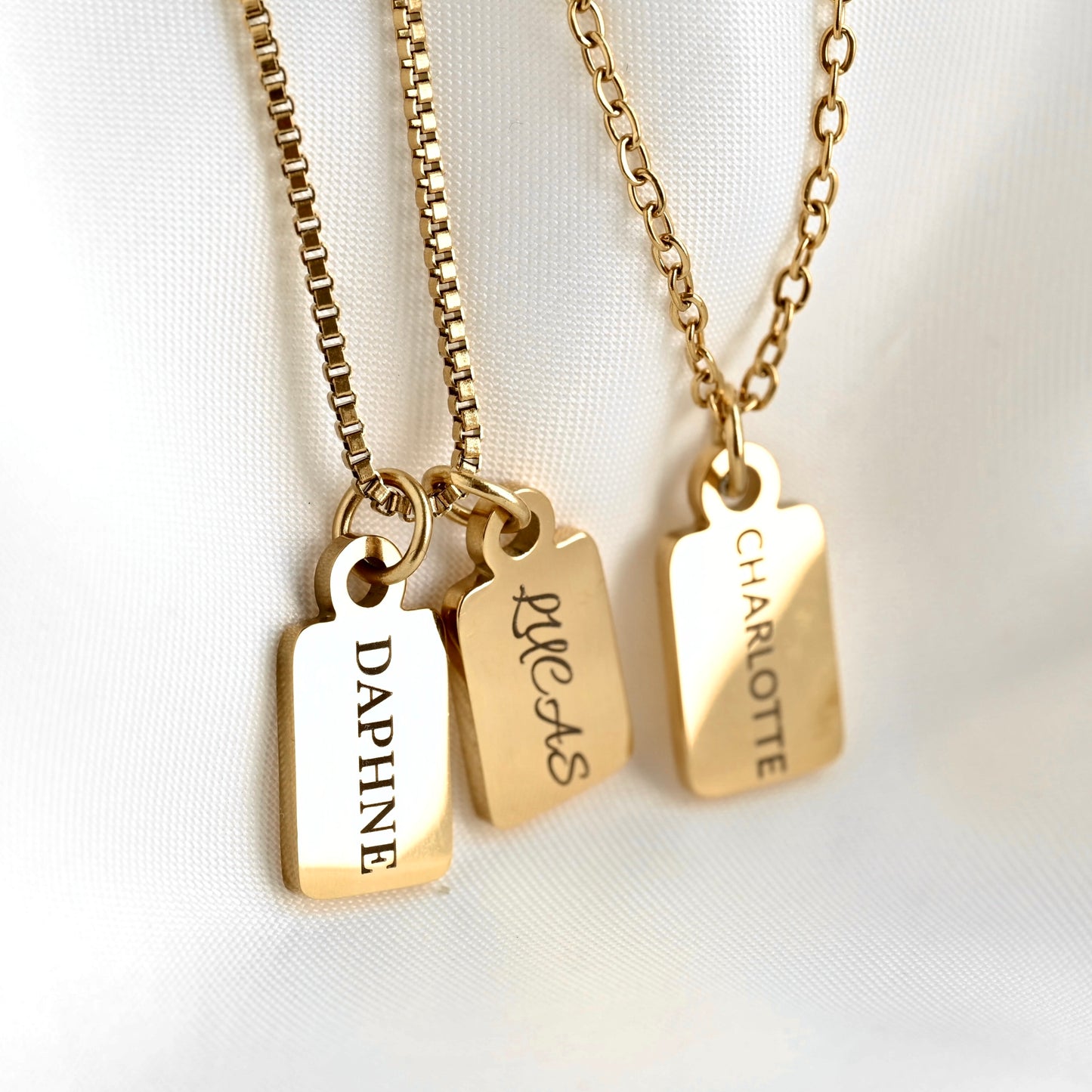Three gold nameplate necklaces with rectangular engraved pendants hanging from box chain and cable chain, displayed on a white fabric background. The pendants are engraved with the names DAPHNE, LUCAS, and CHARLOTTE.