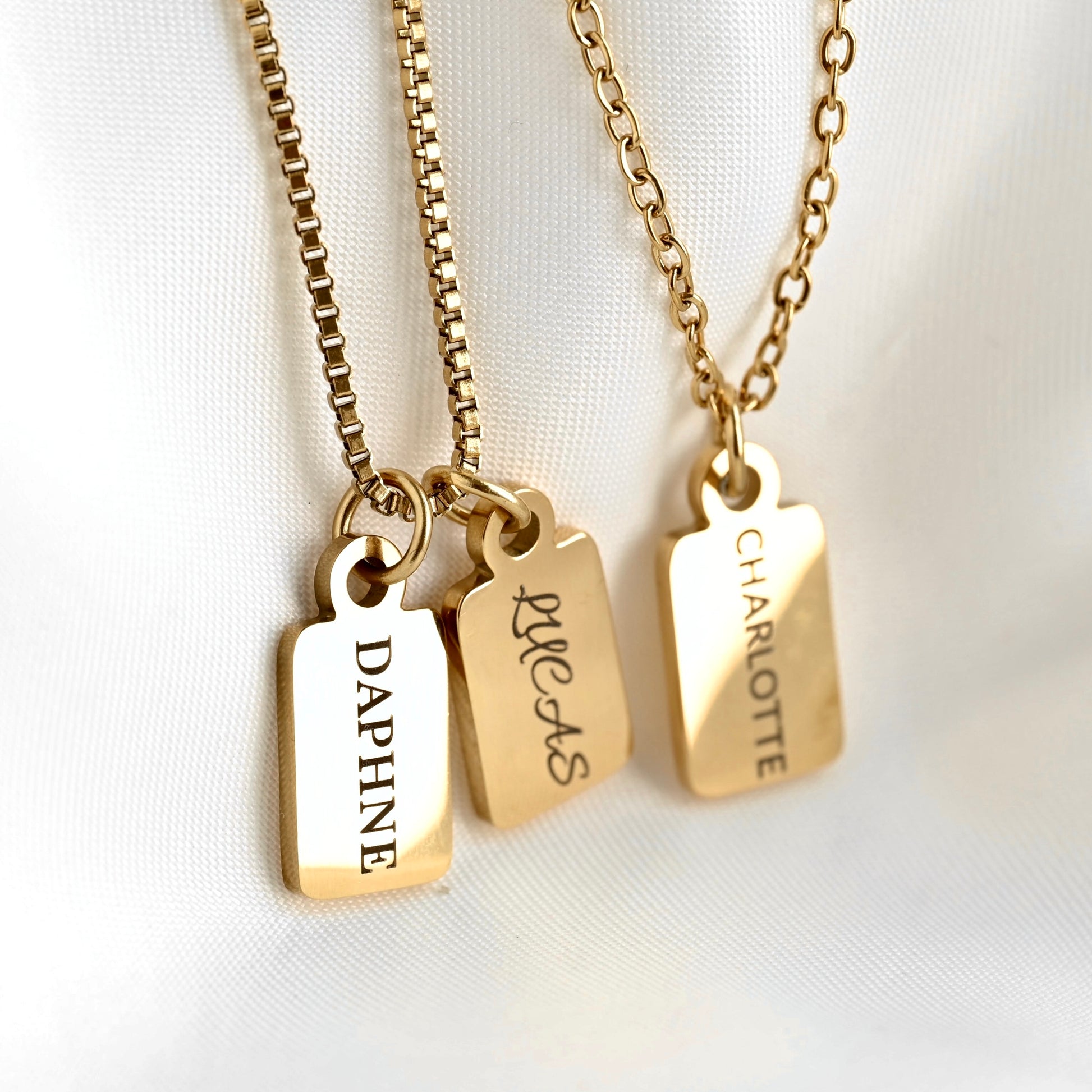 Three gold nameplate necklaces with rectangular engraved pendants hanging from box chain and cable chain, displayed on a white fabric background. The pendants are engraved with the names DAPHNE, LUCAS, and CHARLOTTE.