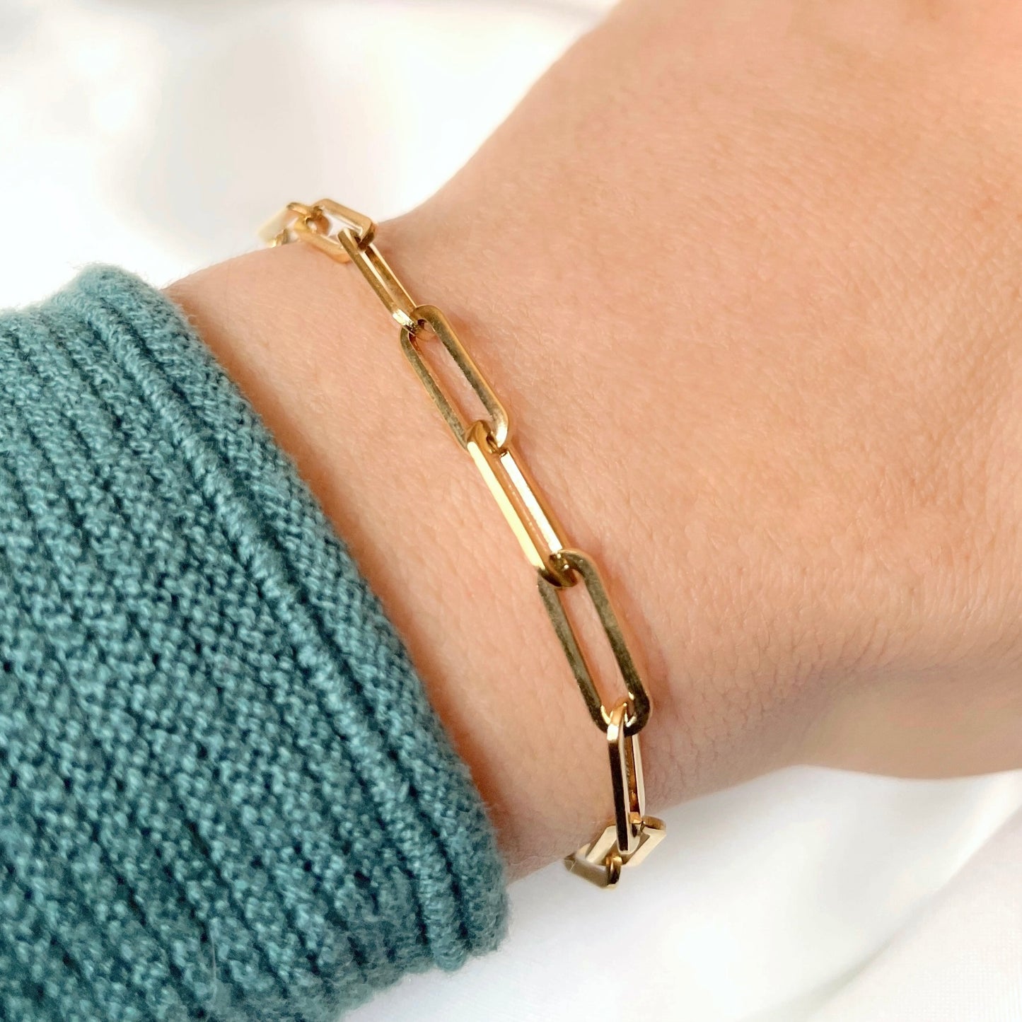 Gold chunky paperclip chain bracelet on a wrist with a green sweater sleeve against a white background