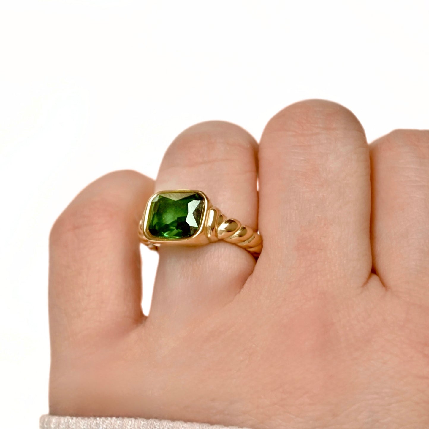 A gold ring with a square-cut emerald gemstone and twisted band, shown worn on a person's finger against a plain white background.