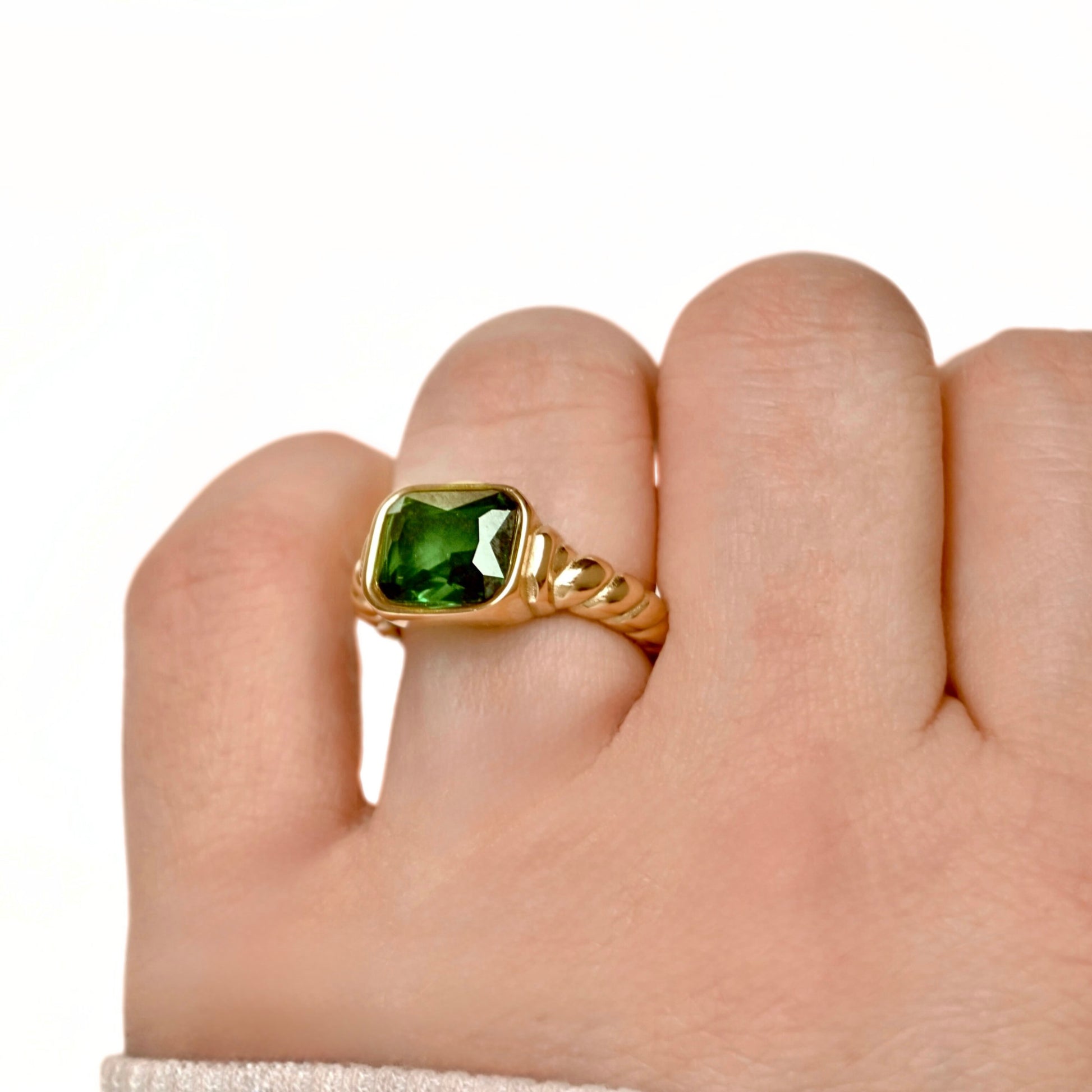 A gold ring with a square-cut emerald gemstone and twisted band, shown worn on a person's finger against a plain white background.