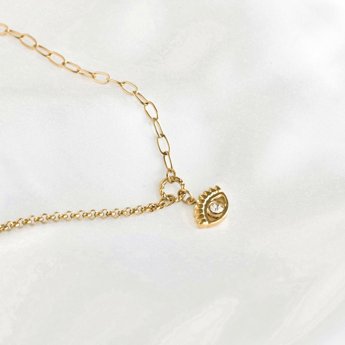 A gold paperclip chain necklace with an evil eye charm featuring a clear crystal, displayed on a soft white fabric background.