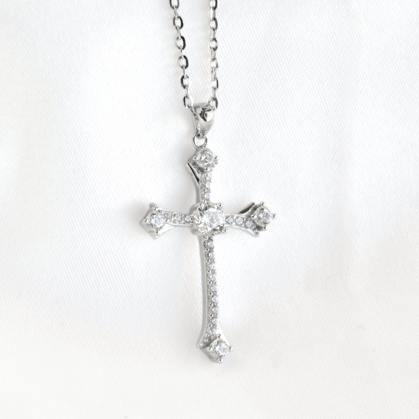 A silver cross pendant necklace adorned with clear gemstones, hanging from a silver chain against a white fabric background.