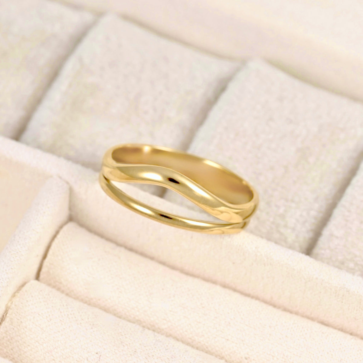 A polished gold ring with a wavy, double-band design resting on a textured cream-colored jewelry display box.