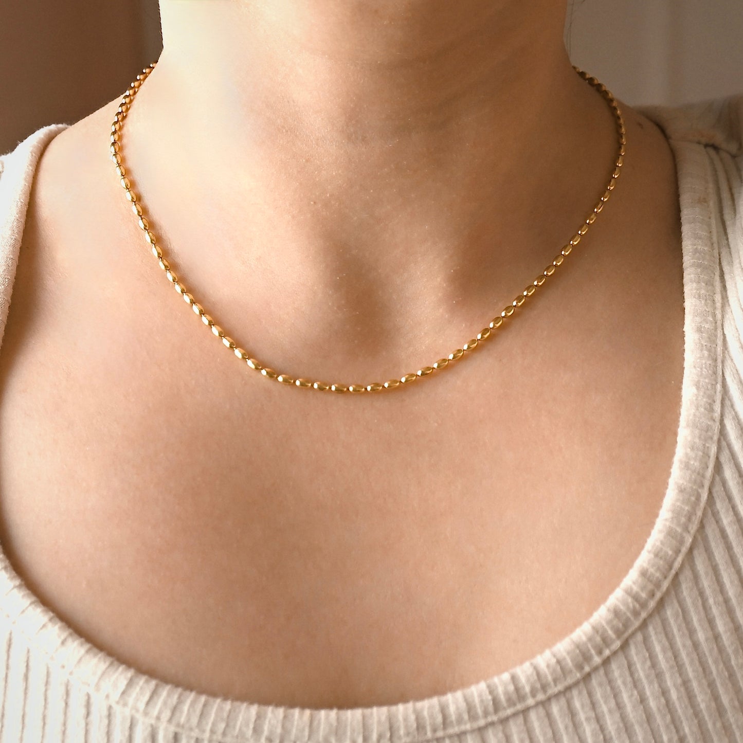 A delicate gold beaded chain necklace worn by a person, shown in a close-up shot of the neck and upper chest wearing a cream-colored ribbed top.