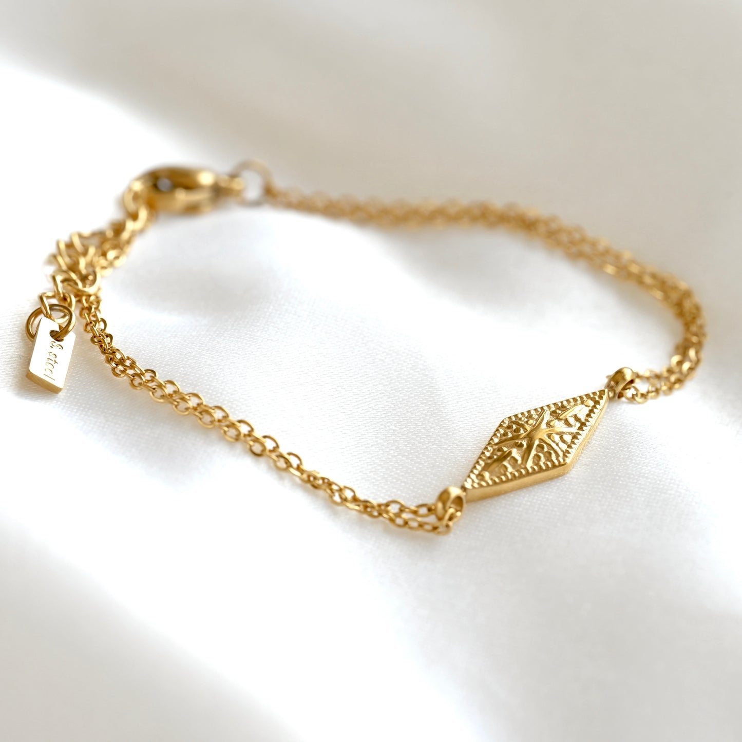 A delicate gold chain bracelet with a textured diamond-shaped centerpiece with a sunburst design, displayed on a soft white fabric background.