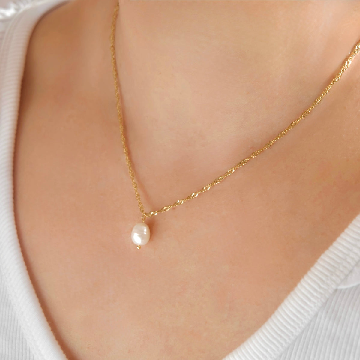 A delicate gold chain necklace with a single small white pearl pendant, worn on a model's neck over a white ribbed top.