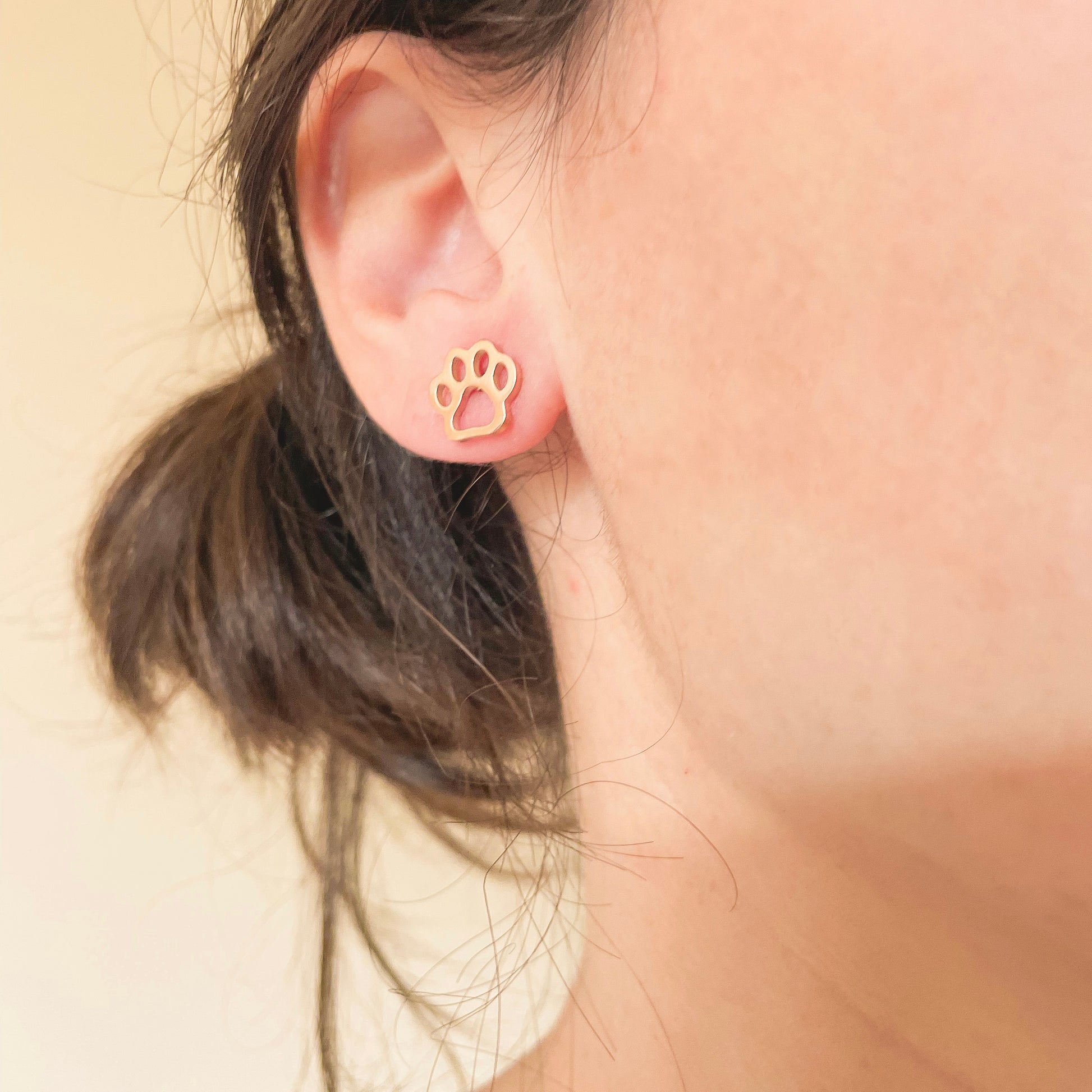Rose gold paw print earrings worn by a person on a beige background