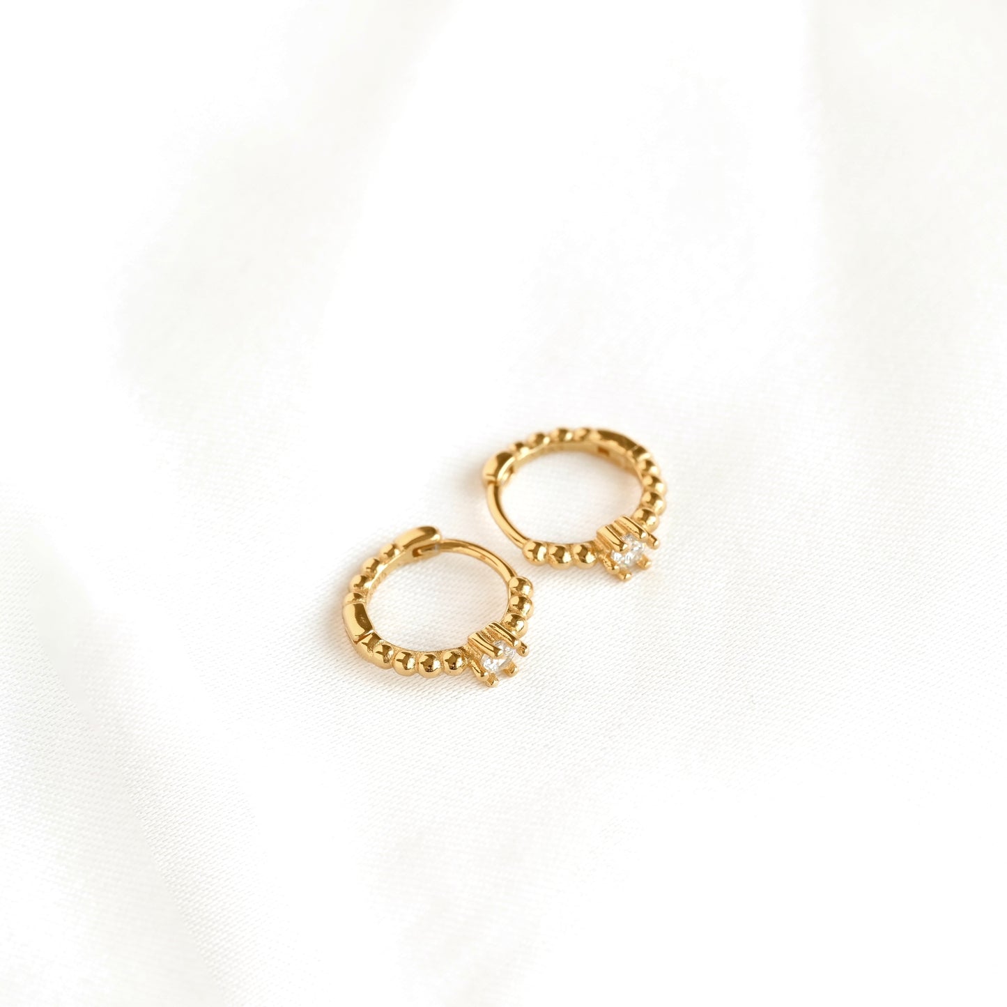Top view of a pair of delicate gold huggie hoop earrings with textured beaded design and small clear crystal accents, displayed on a soft white fabric background.