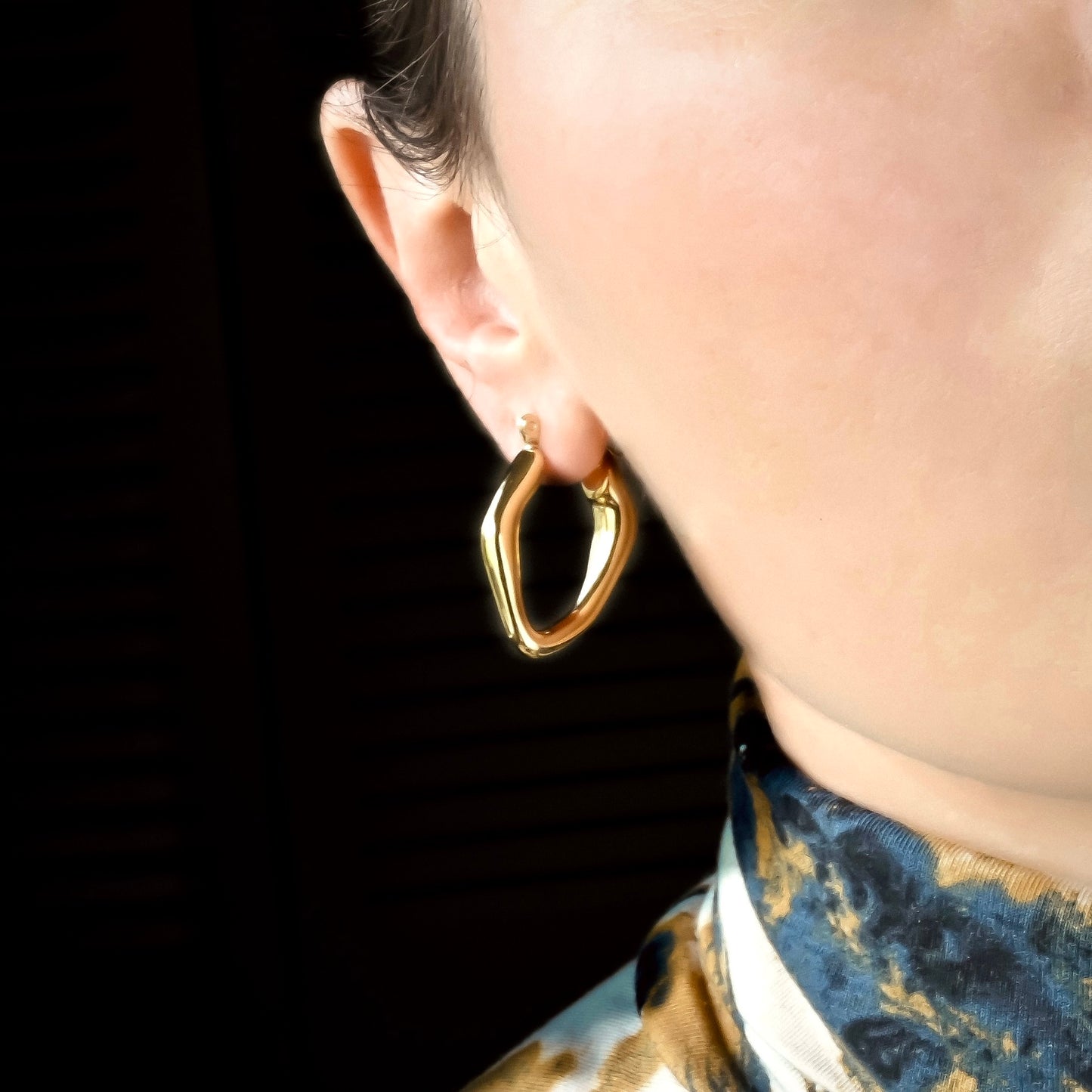 Thin gold scultural hoop earrings worn by a person against a dark background