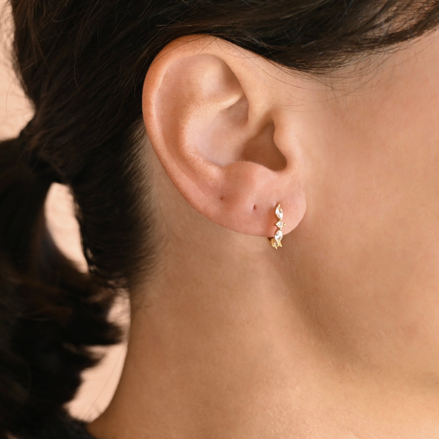 A close-up of a person's ear wearing a small gold huggie earring with a a mix of marquise and roun-cut CZ stones.