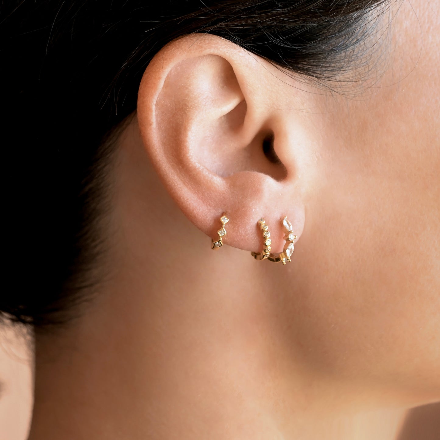 Close-up of a person's ear wearing three small gold huggie hoop earrings, with dark hair visible at the top.