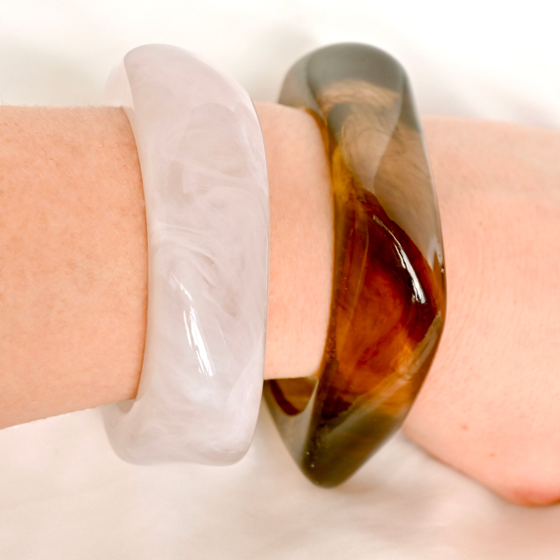 Two translucent bangles worn on a wrist: one milky white with soft marbling, and one dark amber-brown with glossy tiger-stripe bands, against a soft white background.