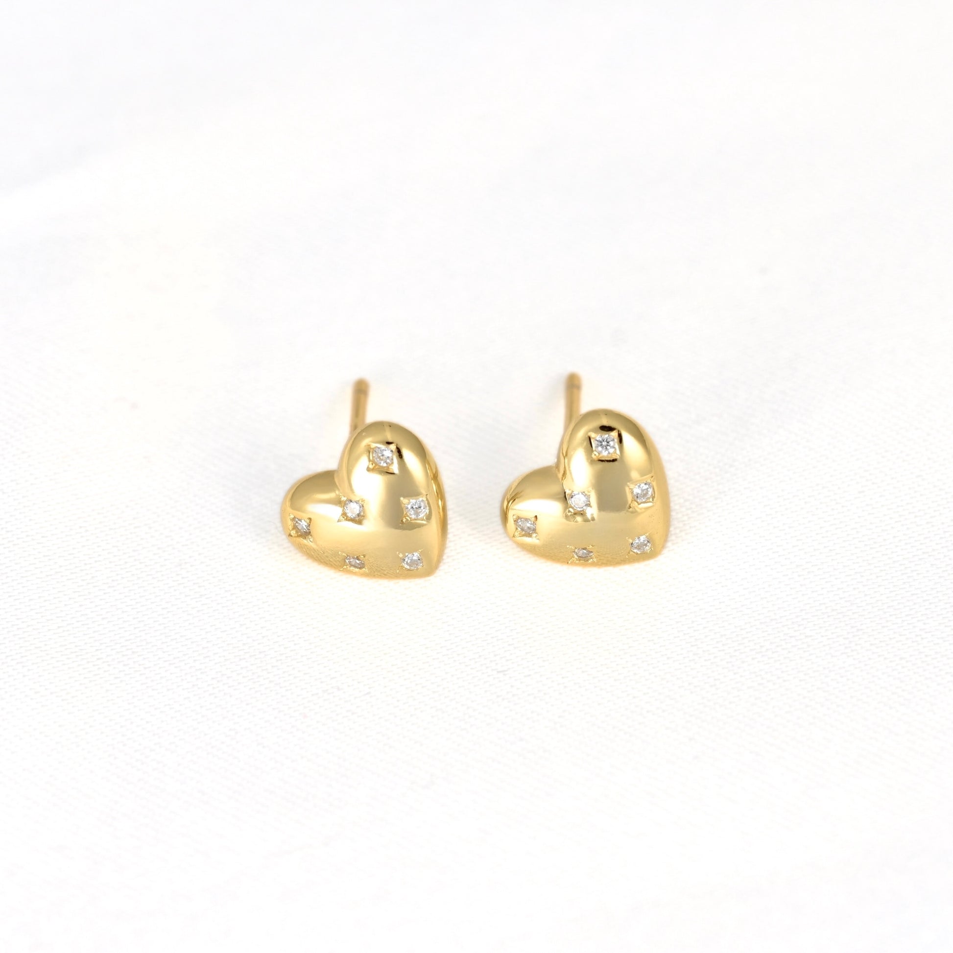 A pair of gold heart stud earrings with small clear crystal accents, displayed on a clean white textured background.