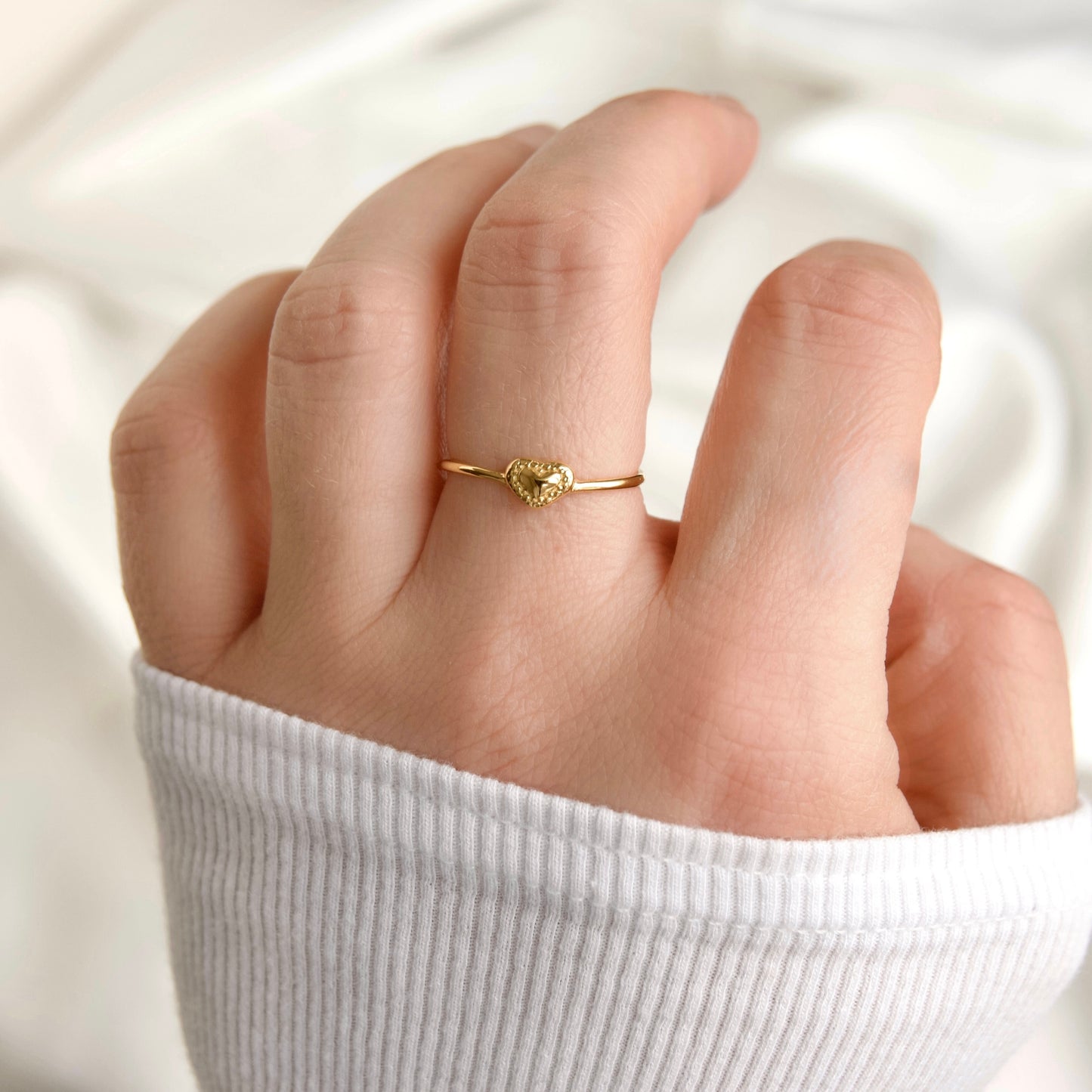 A delicate gold ring with a small textured heart charm, worn on a person's ring finger. The hand is resting on a soft, white ribbed fabric sleeve, set against a soft, out-of-focus white background.