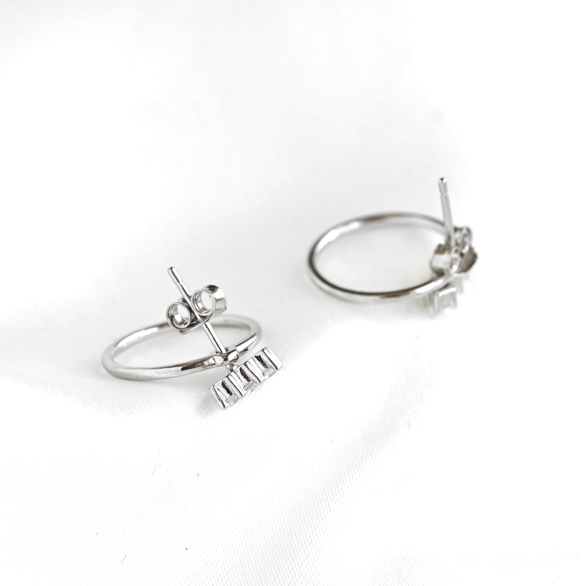A pair of silver ear jacket earrings with a small bar with three clear stones on the front and a polished circle at the back, photographed on a white fabric background.