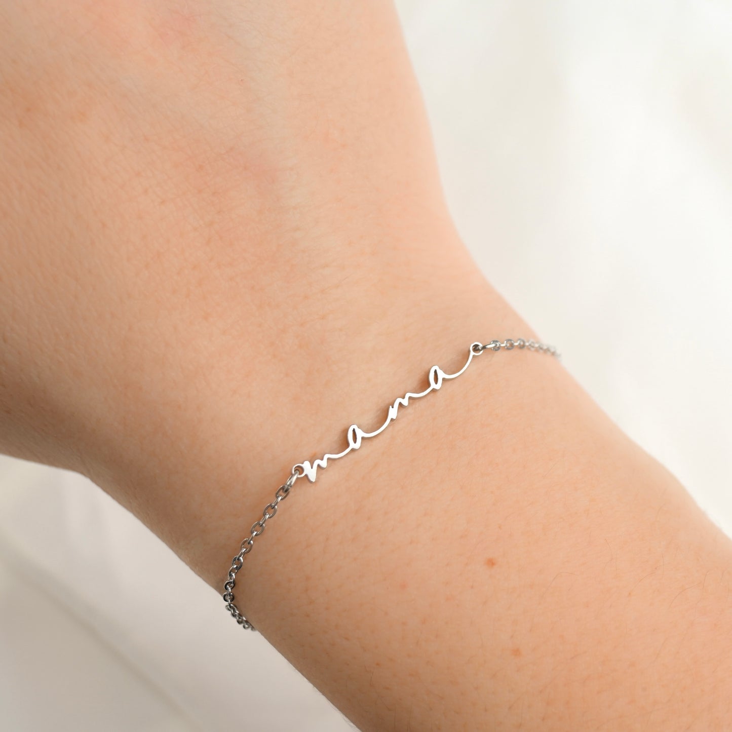 A delicate silver chain bracelet with a cursive 'mama' nameplate, shown worn on a person's wrist against a soft white background.