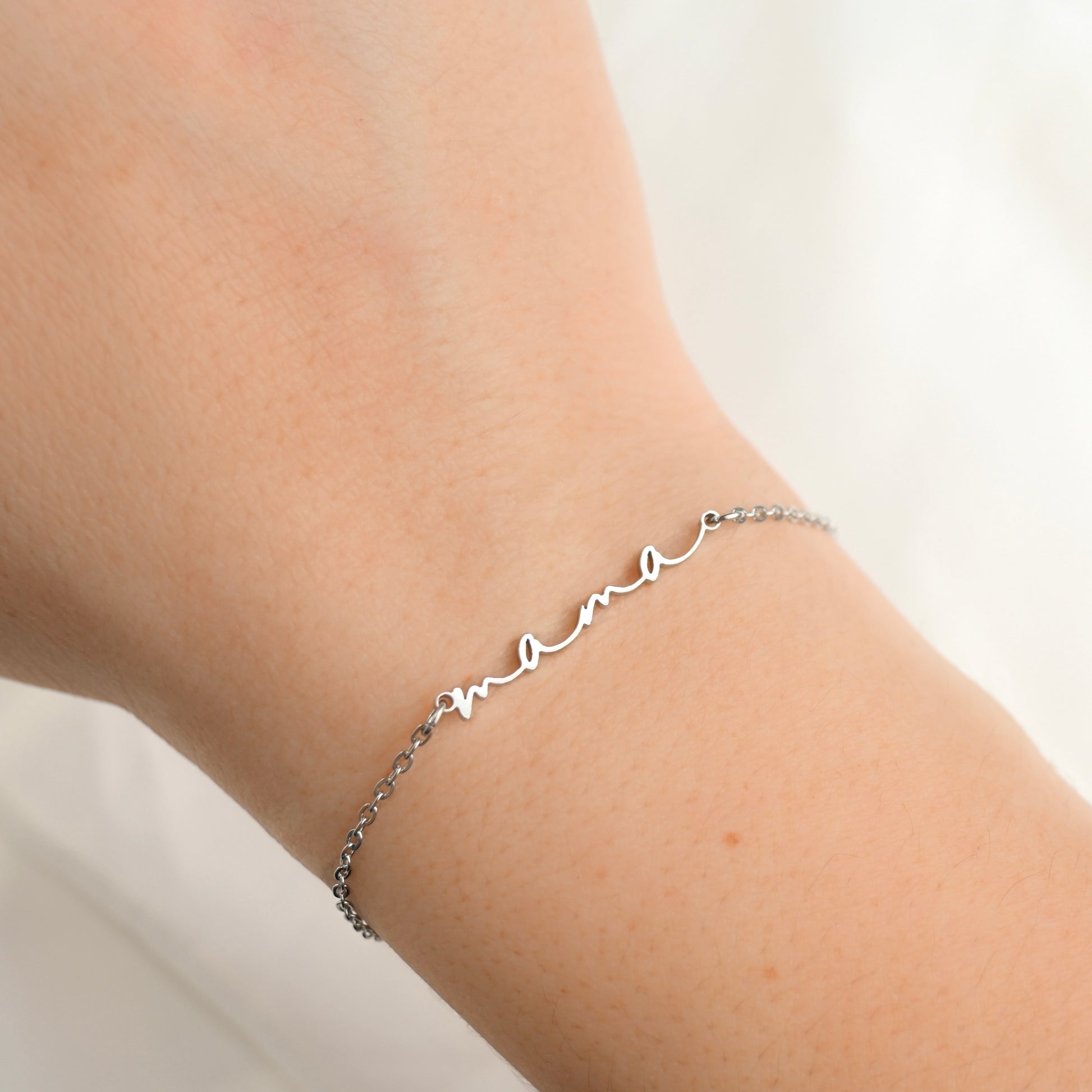 A delicate silver chain bracelet with a cursive 'mama' nameplate, shown worn on a person's wrist against a soft white background.