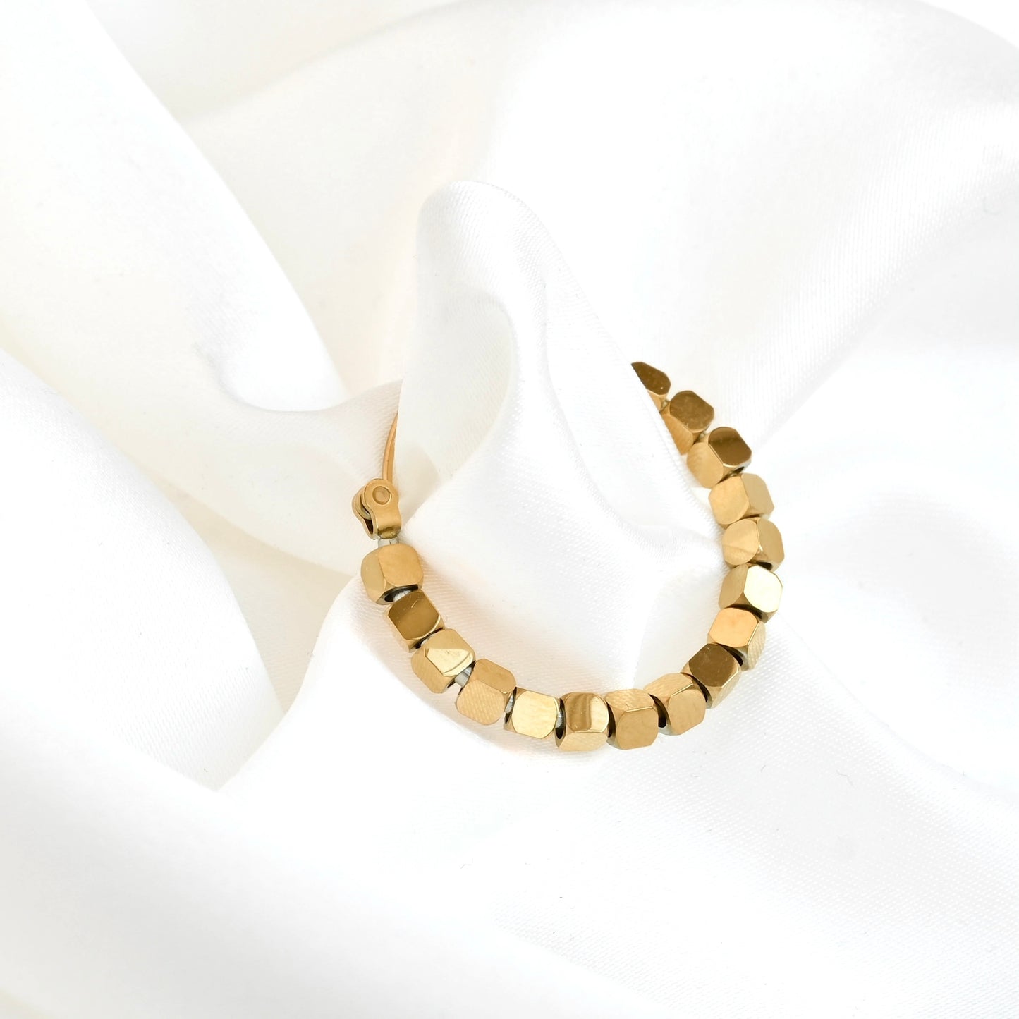 A gold hoop earring with faceted, geometric cube beads, displayed on a soft, draped white fabric background.