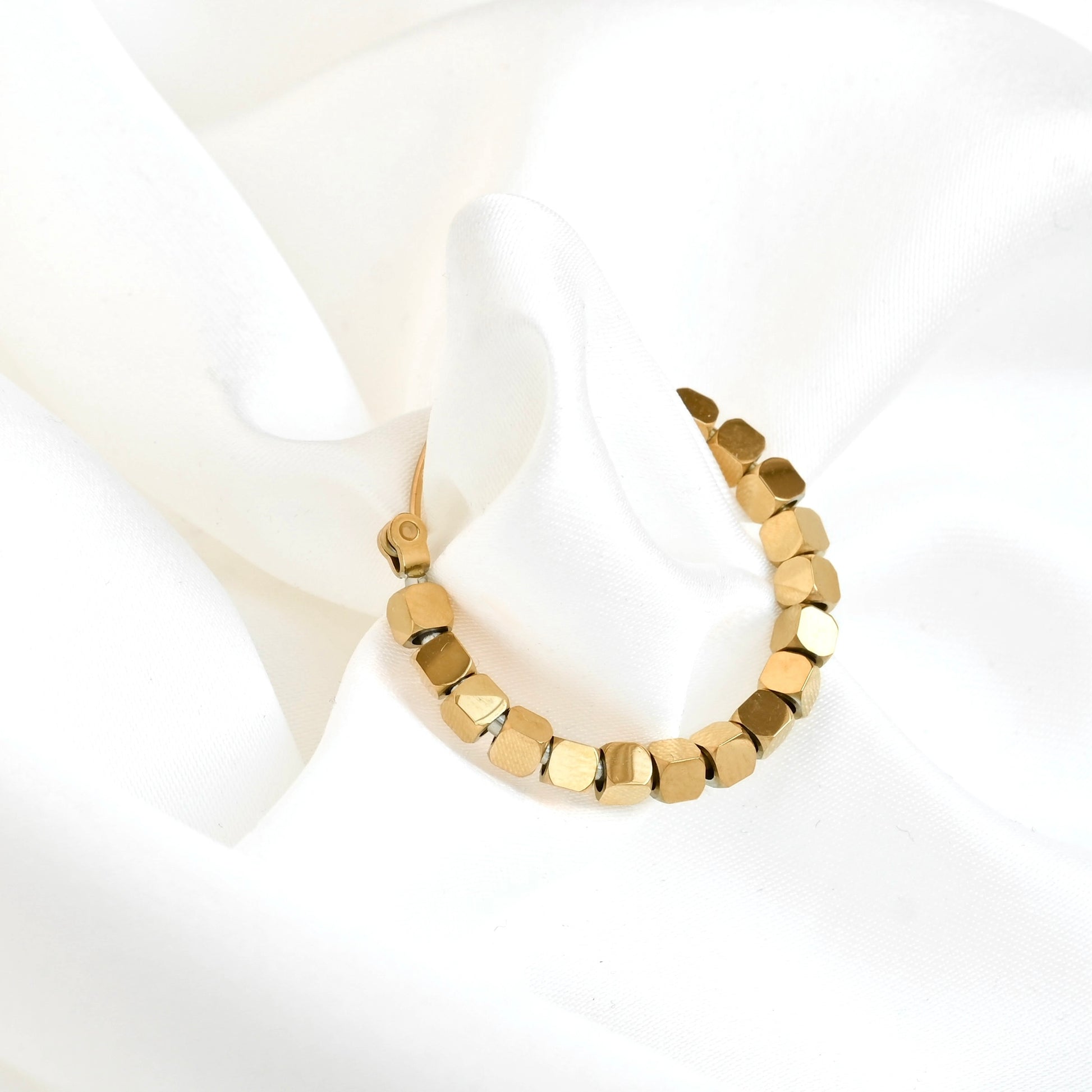 A gold hoop earring with faceted, geometric cube beads, displayed on a soft, draped white fabric background.