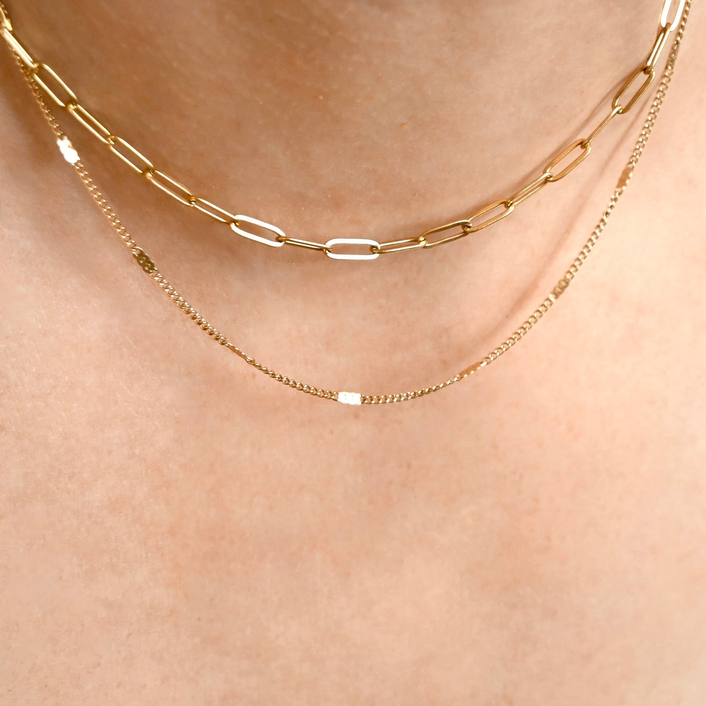 Two layered gold chain necklaces worn on a person's neck: a thicker paperclip chain and a delicate chain with small rectangular links.