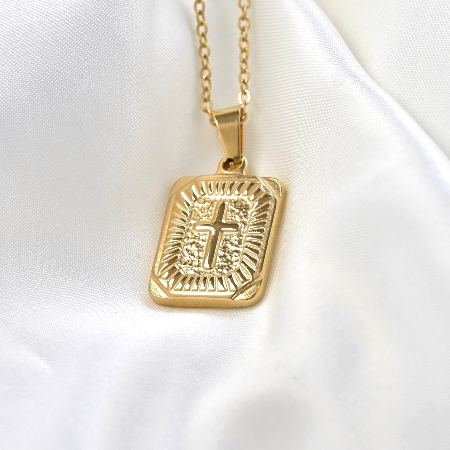 A gold-toned rectangular pendant necklace with an embossed cross and decorative border, hanging from a matching chain on a white satin fabric background.
