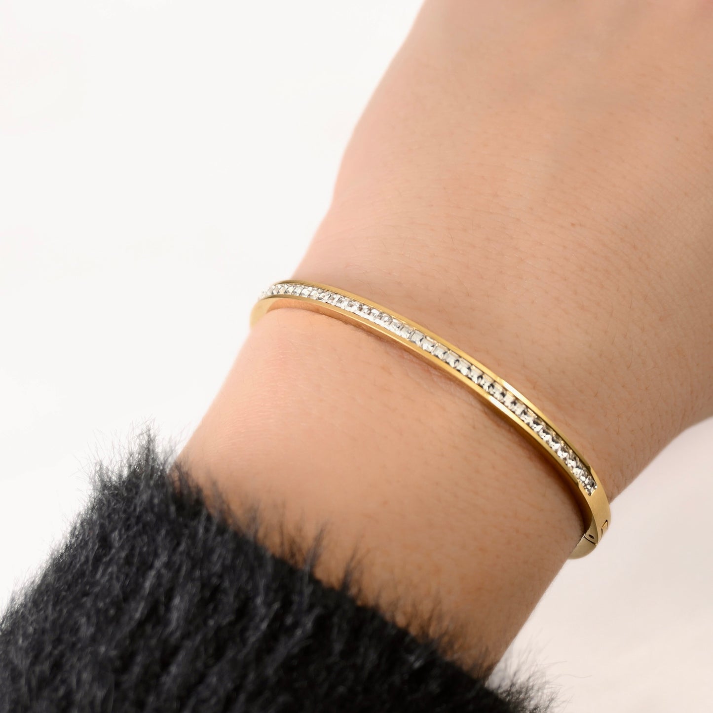 A gold bangle bracelet with a row of clear crystals, worn on a person's wrist against a white background.