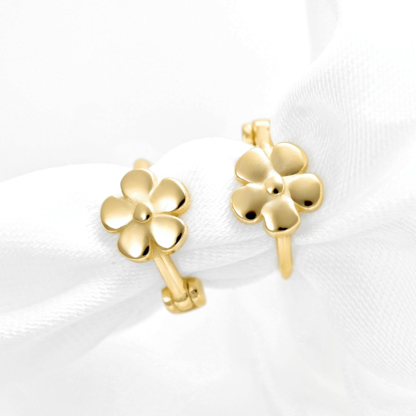 A pair of gold hoop earrings with small five-petal flower charms, displayed on a soft white fabric background.